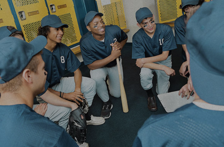 A group of young baseball players in blue uniforms and caps sitting in a locker room, listening to a coach.
