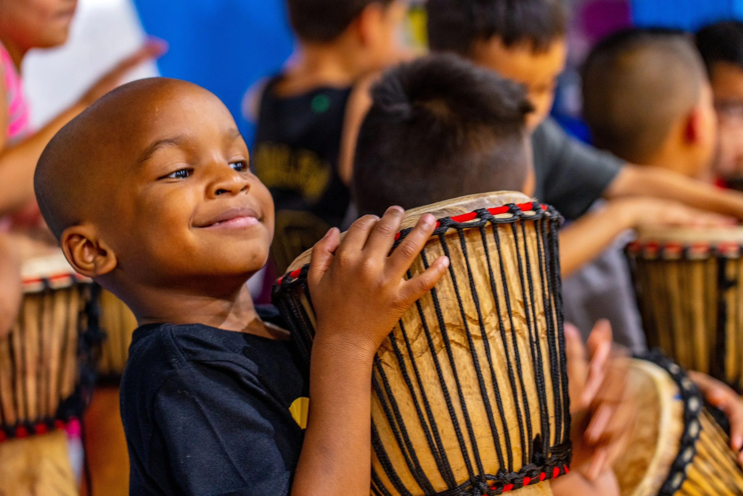 A young boy smiling while playing a wooden drum surrounded by other children also playing drums.