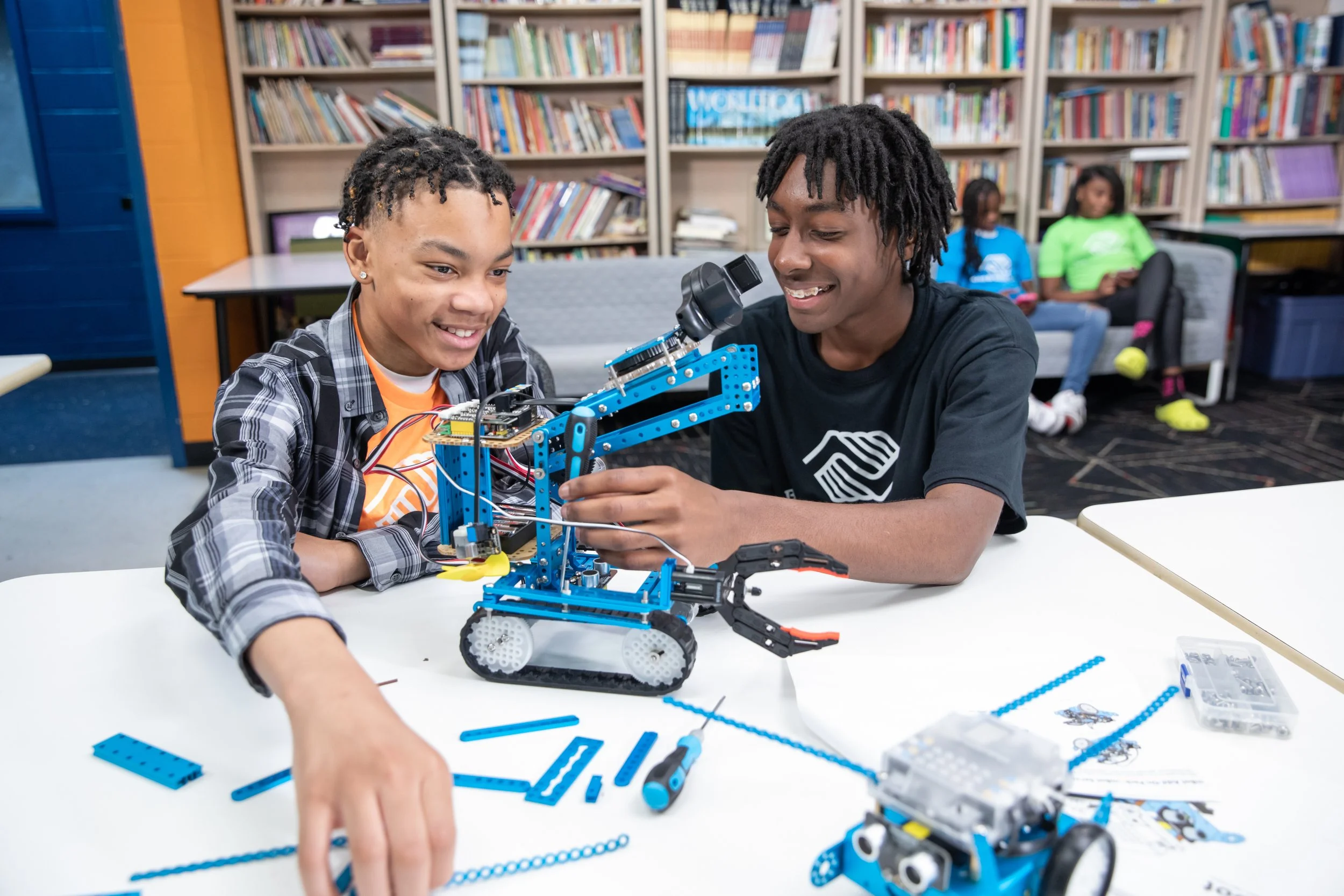 Two boys assembling and working on a blue robotic vehicle at a table in a library or classroom, with two girls sitting on a couch in the background.