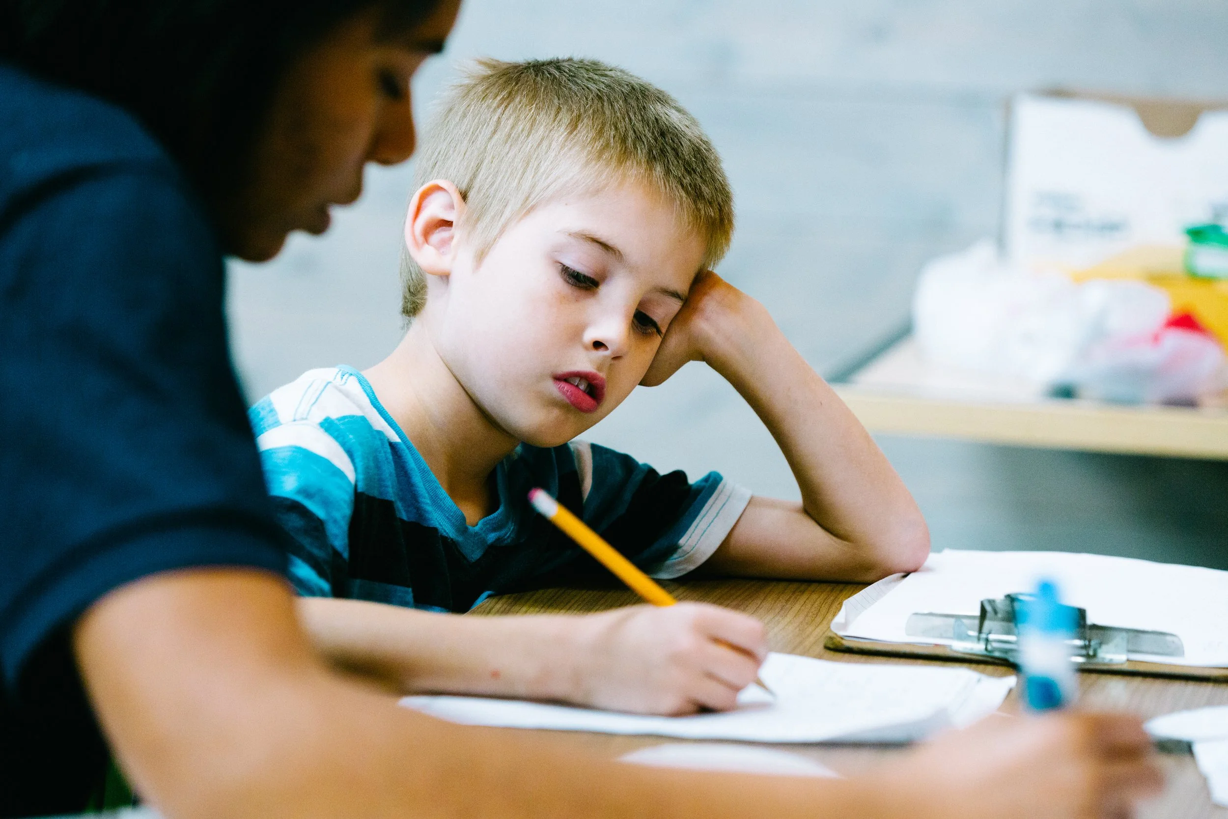 A young boy with blond hair, wearing a striped shirt, sitting at a table, studying with a pencil in his hand, looking focused and pensive.