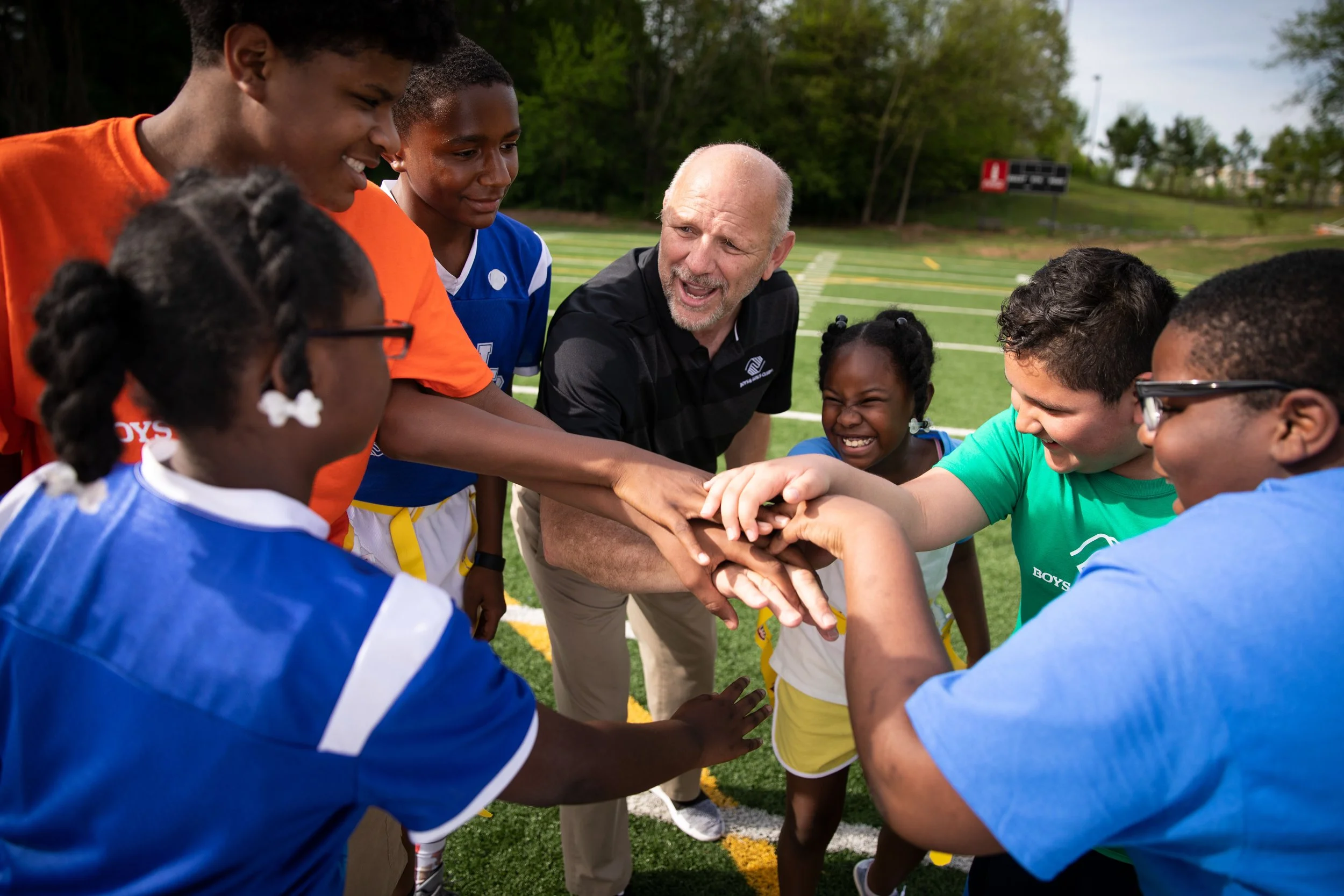 A group of children and an adult coach in a huddle on a sports field, putting their hands together in the center as a team cheer before a game.