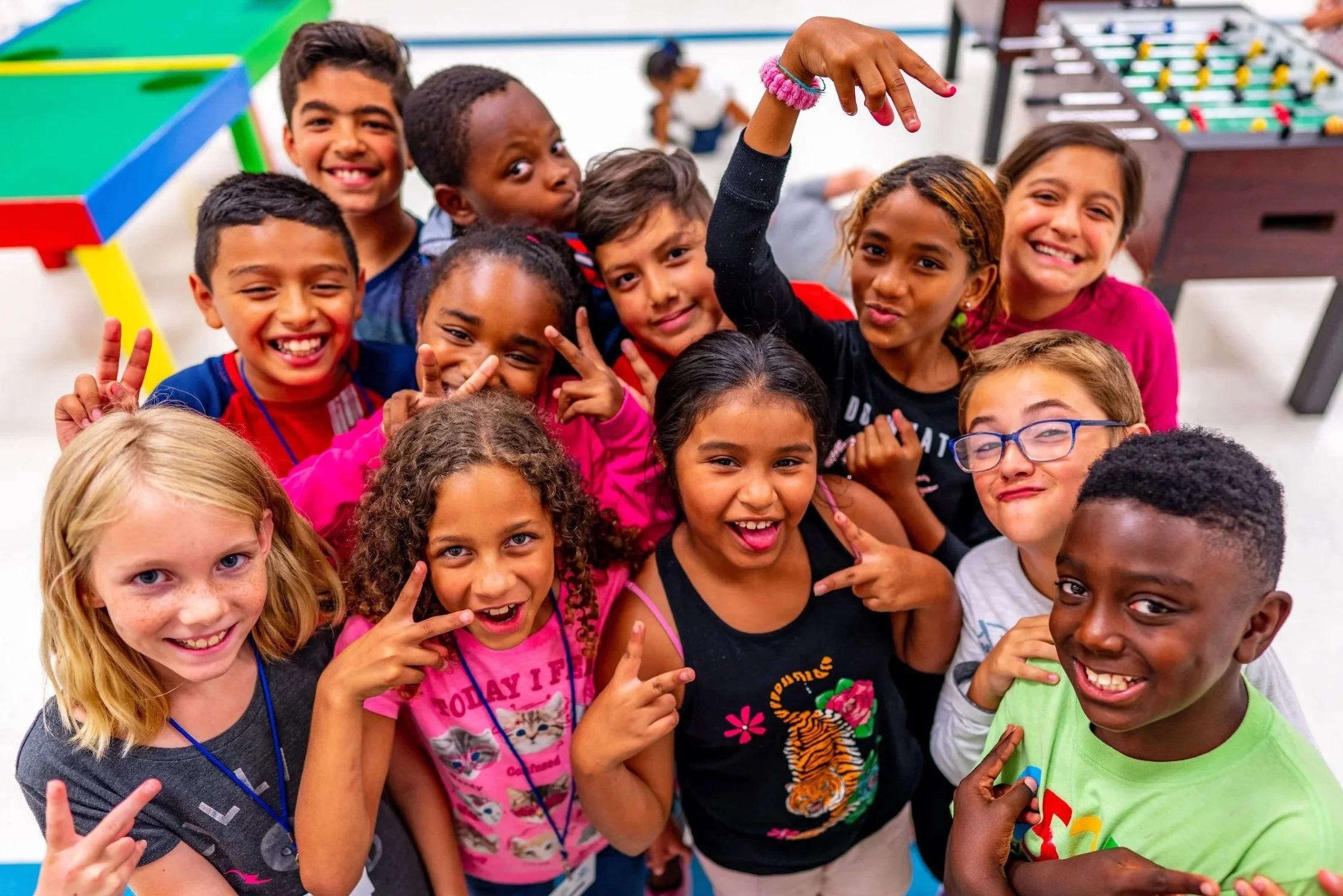 A group of diverse children smiling and making peace signs, gathered in a colorful indoor play area with foosball and game tables in the background.