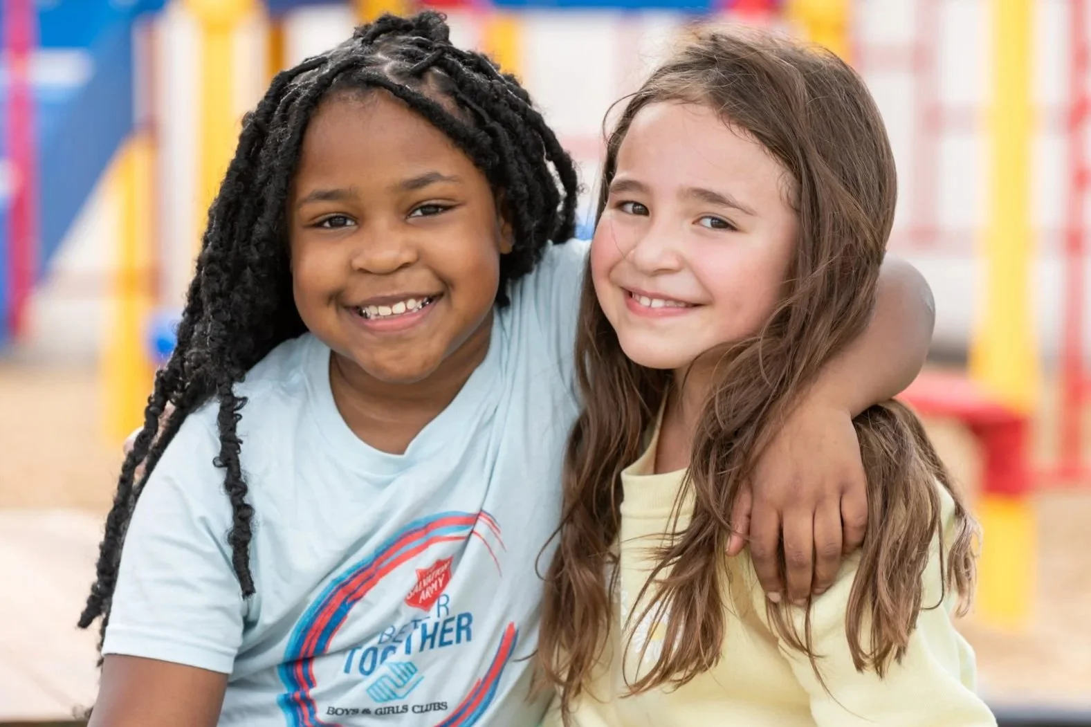 Two smiling young girls with their arms around each other, standing in a brightly colored indoor play area or classroom.