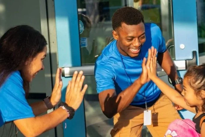 A young man in a blue shirt high-fives a young girl at an entrance of a building, with another woman clapping nearby.