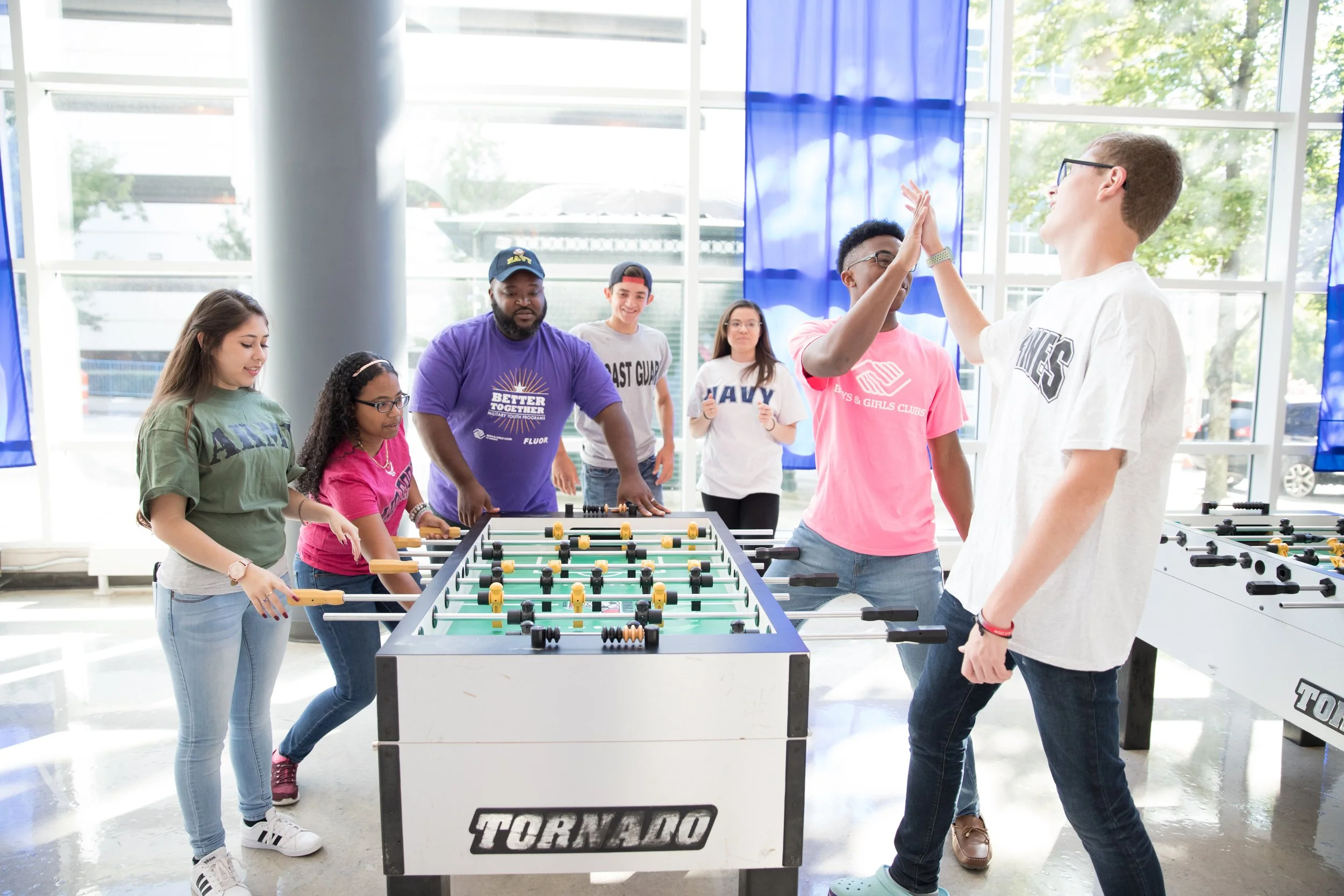 A group of diverse young people playing foosball indoors, with some high-fiving and others watching, near large windows.