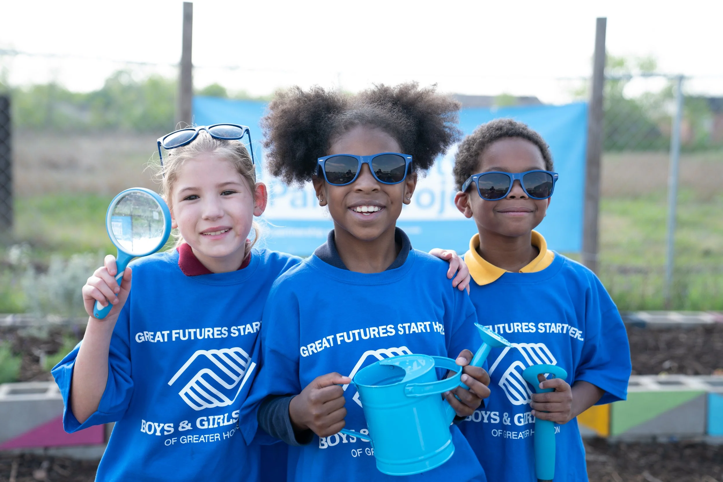 Three smiling children in blue shirts with the phrase 'Great Futures Start Here' and 'Boys & Girls of Greater Houston' printed on them, wearing sunglasses, holding gardening tools and a watering can outdoors.