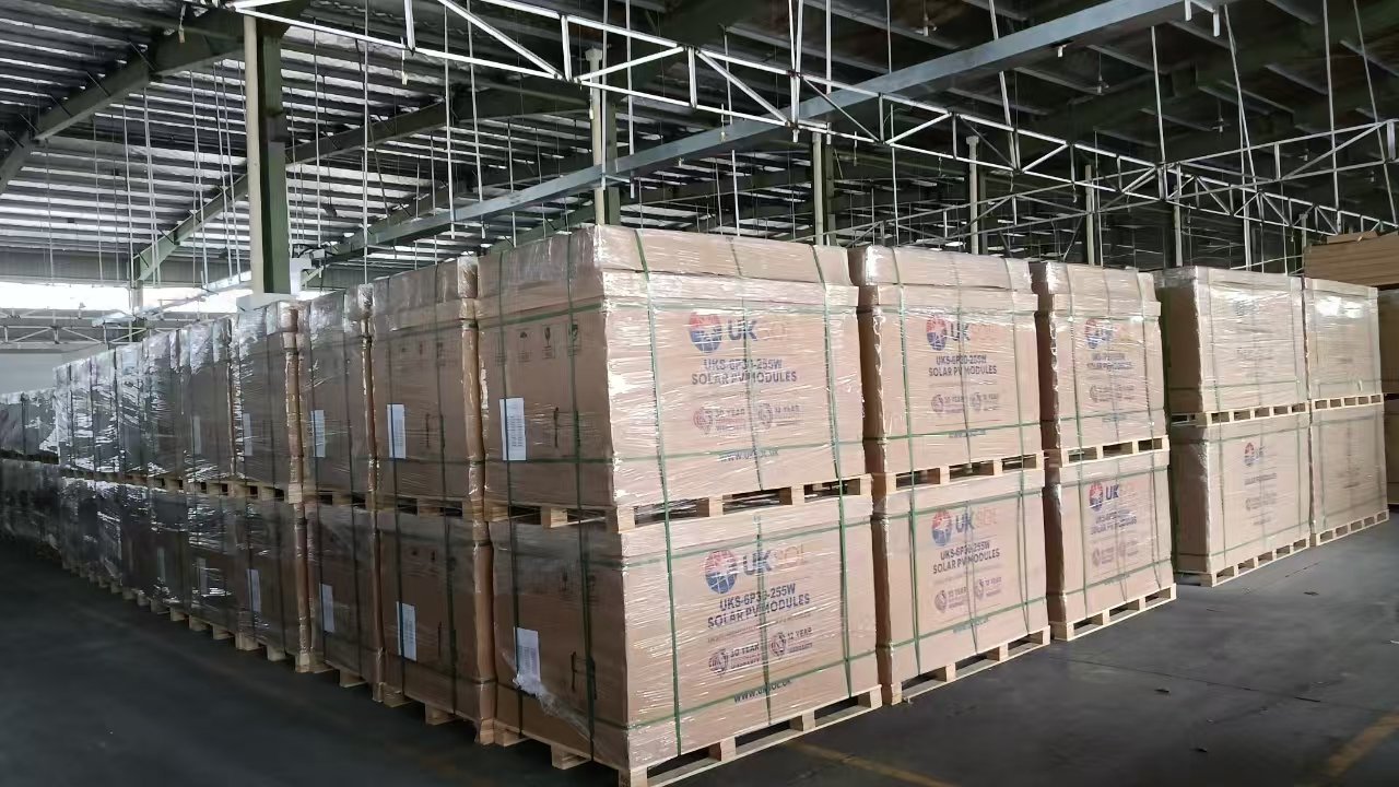 Stacks of boxed solar modules wrapped in plastic and secured with green straps, stored on wooden pallets inside a warehouse.