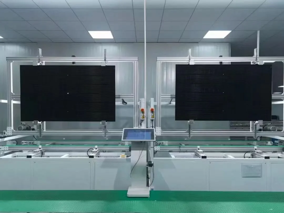 Industrial manufacturing or testing equipment with two large black screens, a control panel on a stand, and mechanical parts in a lab or factory setting.