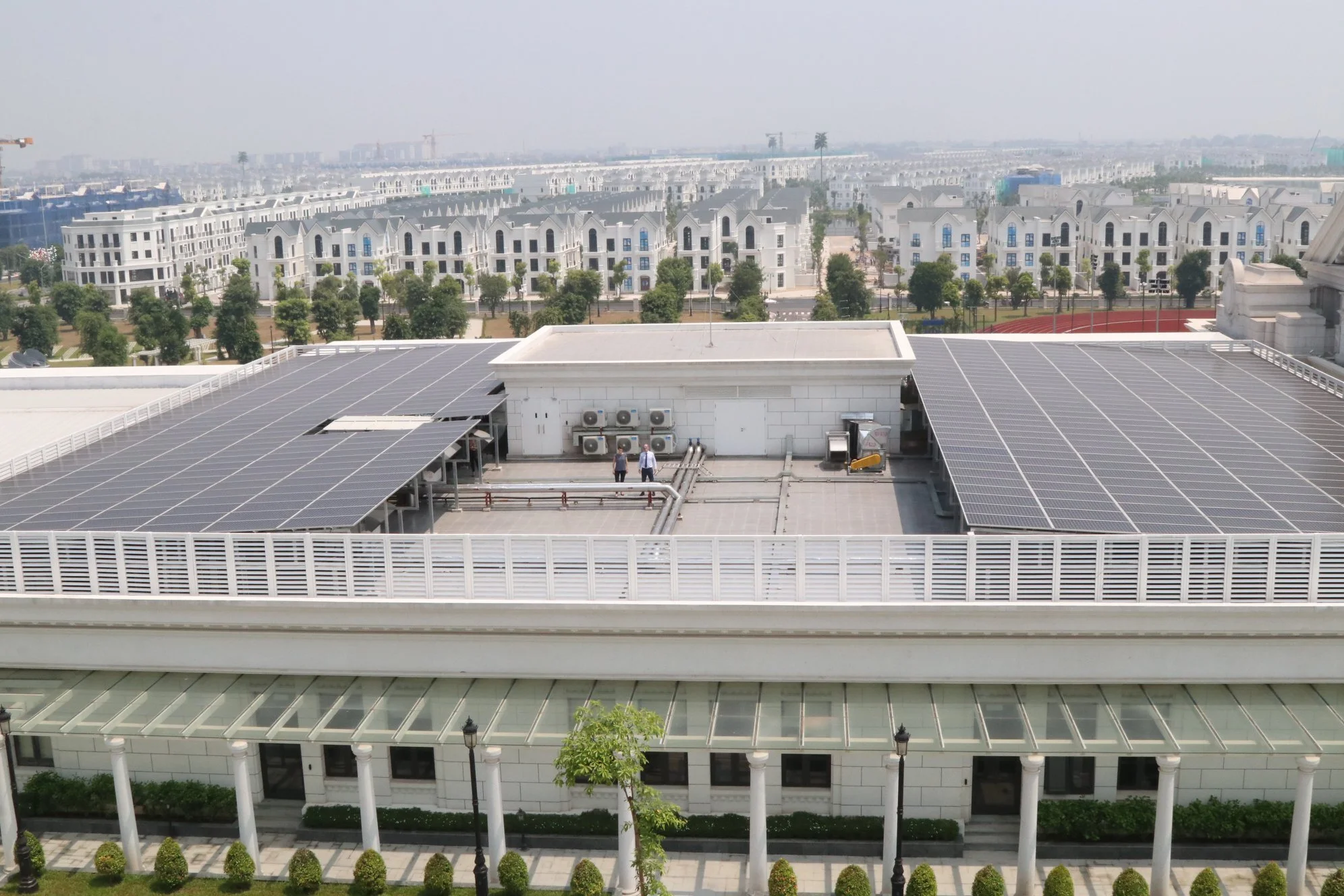 View of a building rooftop with solar panels, air conditioning units, and two people standing near the equipment. In the background are residential buildings and trees.