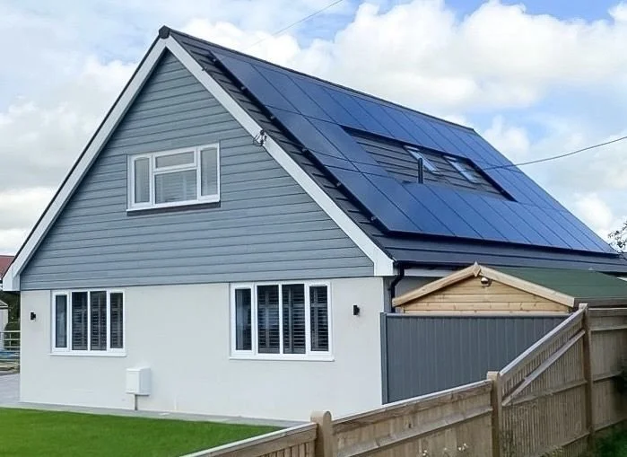 A house with grey siding and white trim, featuring solar panels installed on its steeply angled roof, with a fenced backyard and a partly cloudy sky.