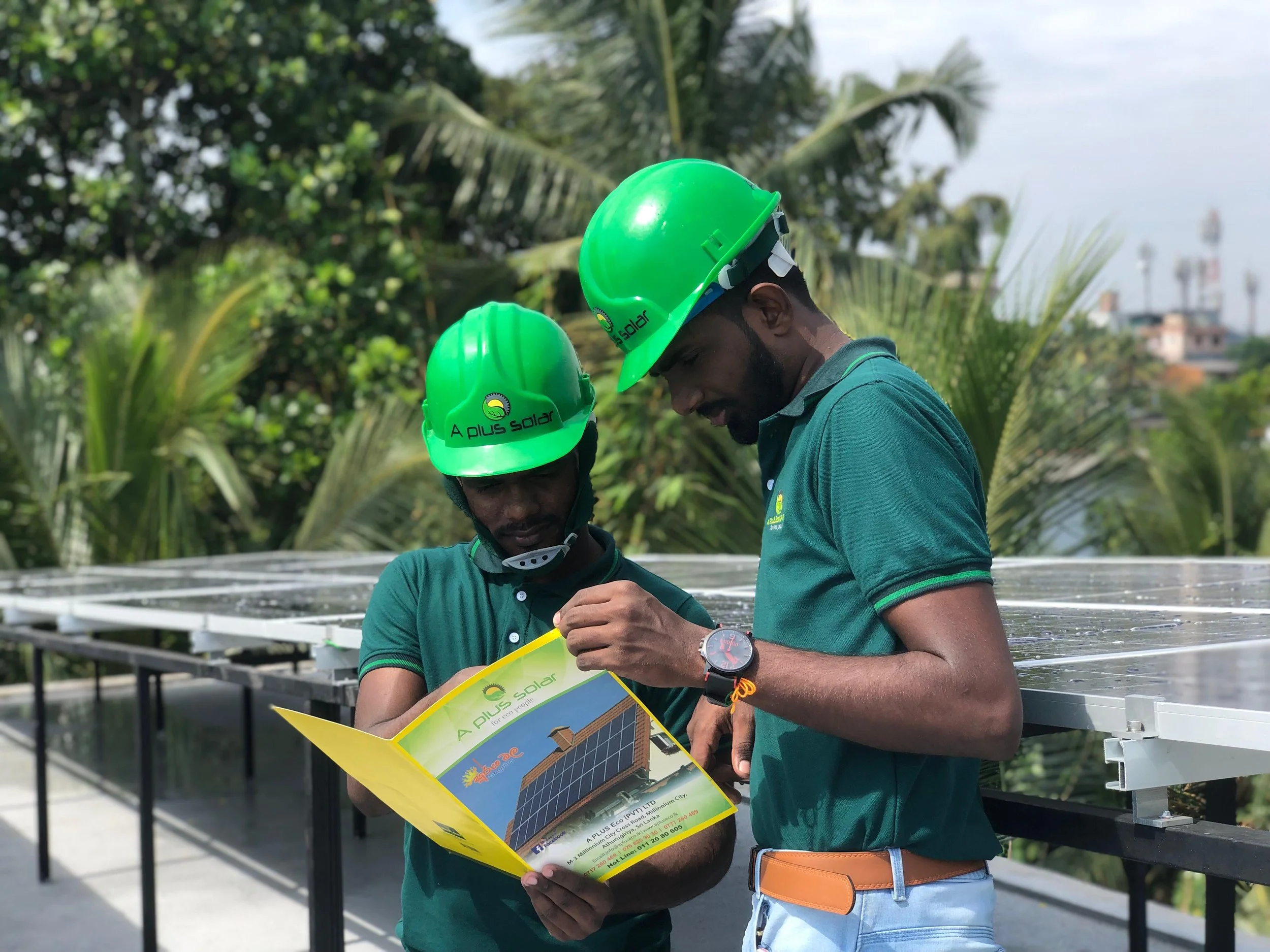 Two workers in green safety helmets and matching green shirts examining a brochure in an outdoor setting with solar panels and trees in the background.