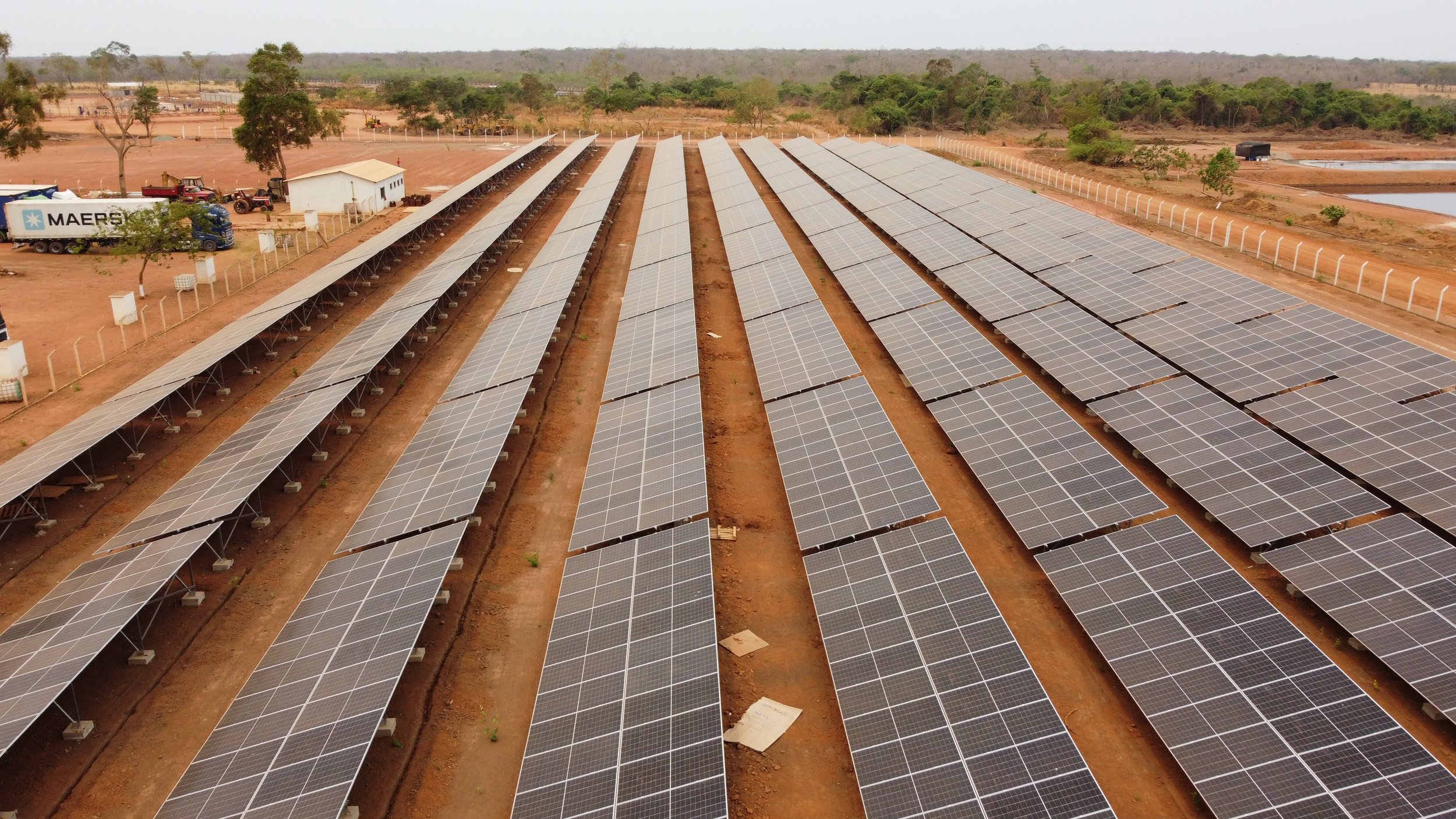 An array of solar panels installed in a large field with dirt pathways between rows, surrounded by a fence, trees, and some construction vehicles nearby.