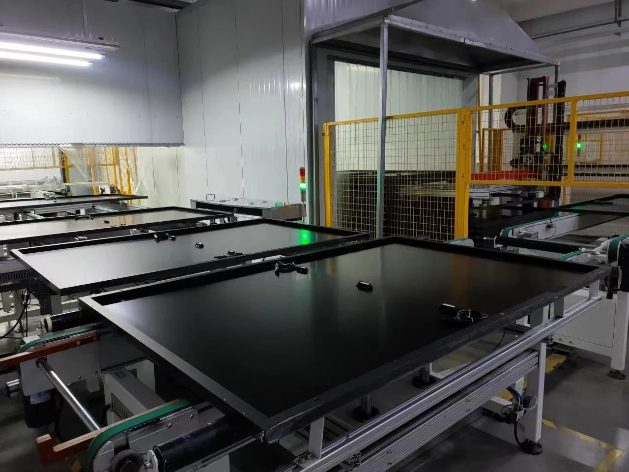 Industrial factory setting with multiple large flat black panels on conveyor belts and a robotic machine enclosed in a yellow safety fence.