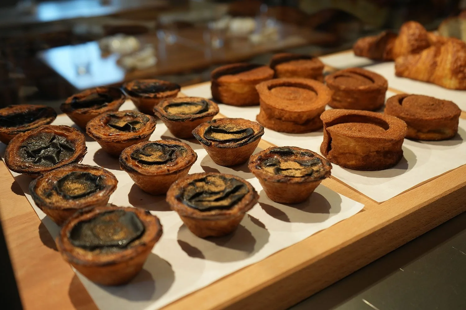 Assorted mini tarts on a wooden display tray, including some with a dark filling and others with a light filling, in a bakery or pastry shop.