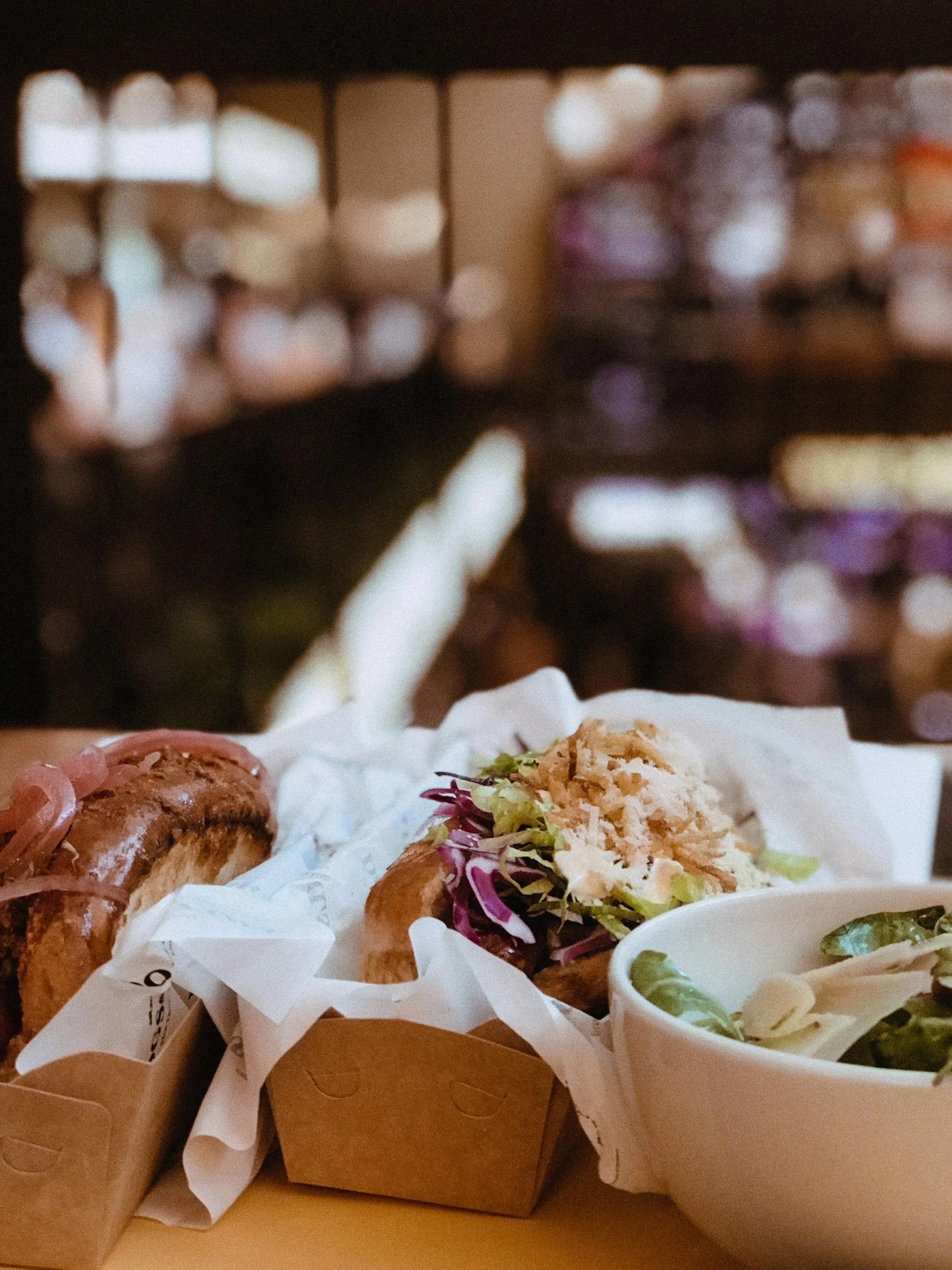 Close-up of two sandwiches with toppings and a bowl of salad on a table, blurred background.