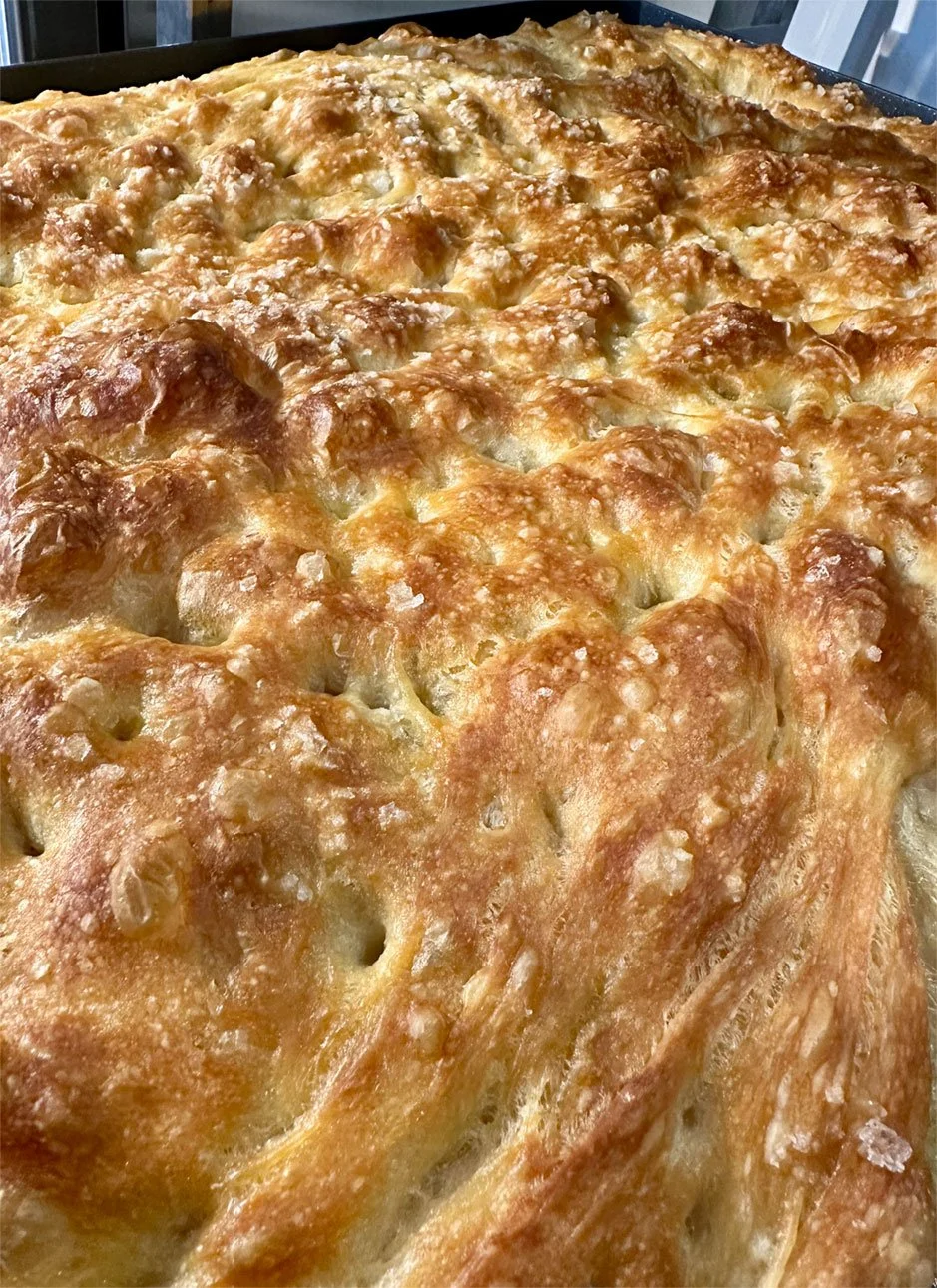 A freshly baked focaccia with a golden brown crispy top, cheese bubbling and slightly browned, in a black baking dish.