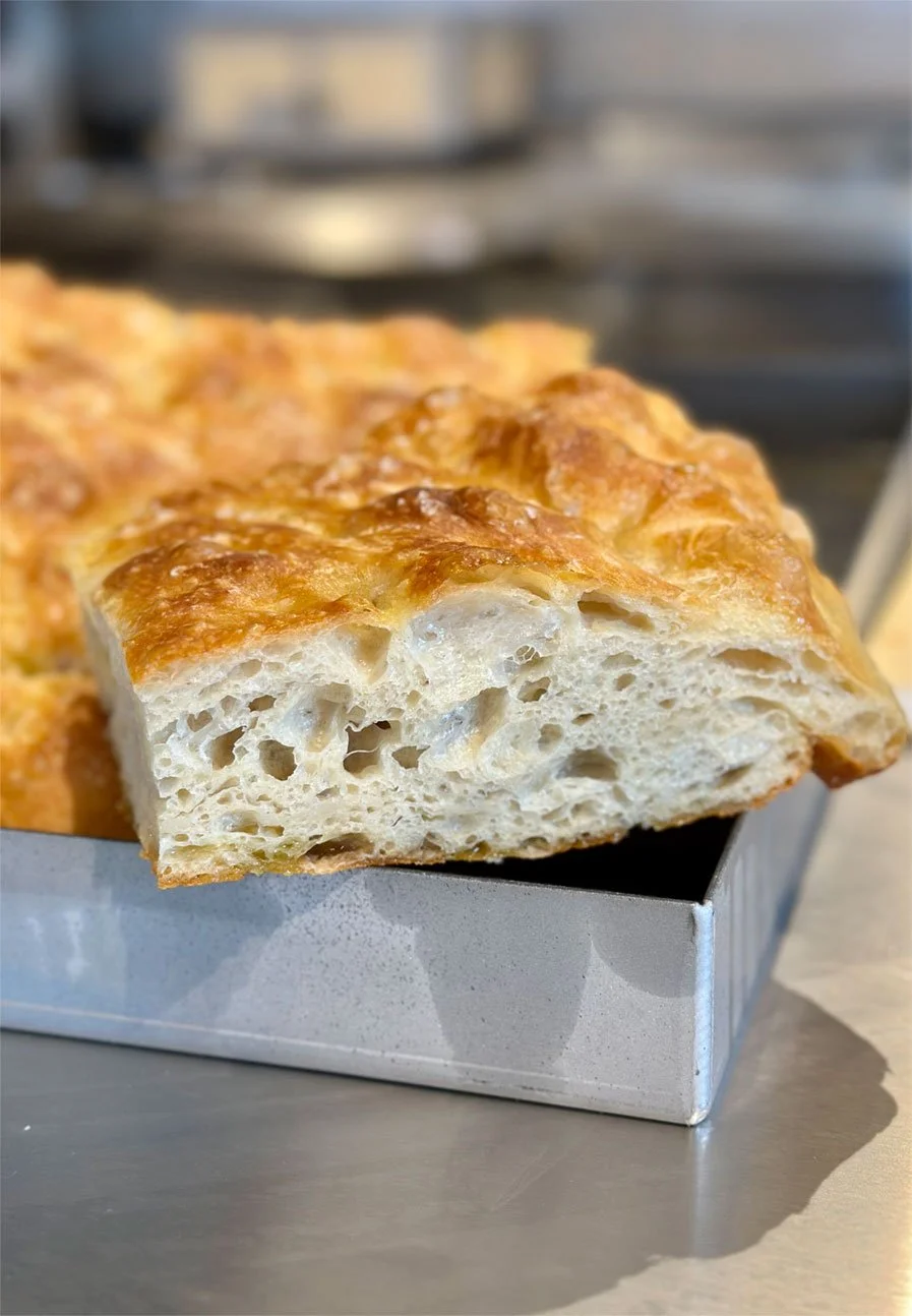 Close-up of a square piece of focaccia bread with a golden, crispy crust and airy, fluffy interior, placed on the edge of a metallic box.
