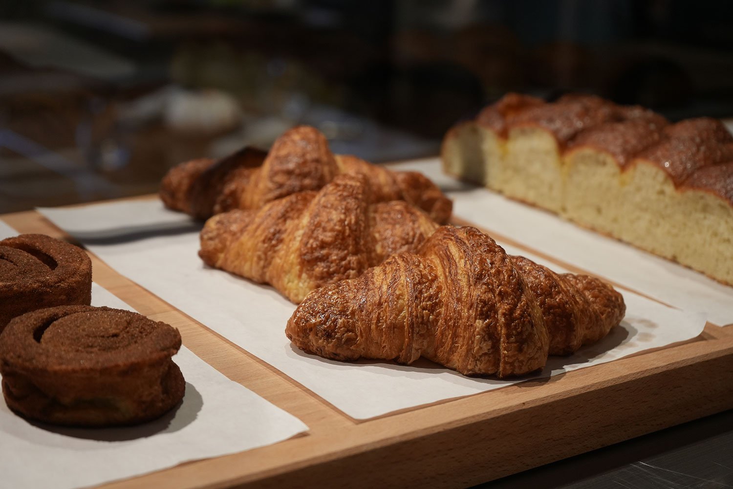 Assorted baked goods on a wooden tray, including croissants, cinnamon rolls, and a slice of cake.