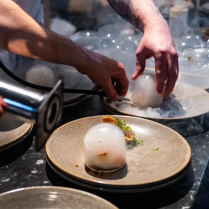 Chef carefully places a spherical food item on a plate surrounded by some garnishes, with another similar spherical item being prepared in a separate dish with dry ice vapor surrounding it.