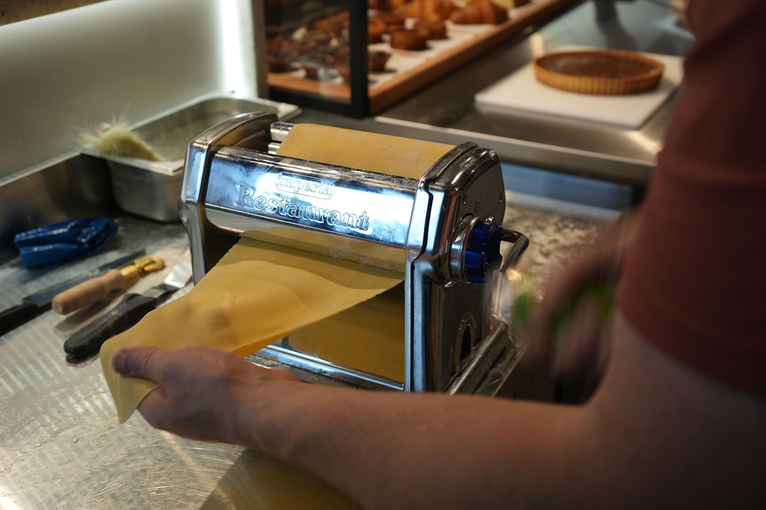 Person using a pasta machine to roll out pasta dough at a kitchen counter, with tools and ingredients nearby.