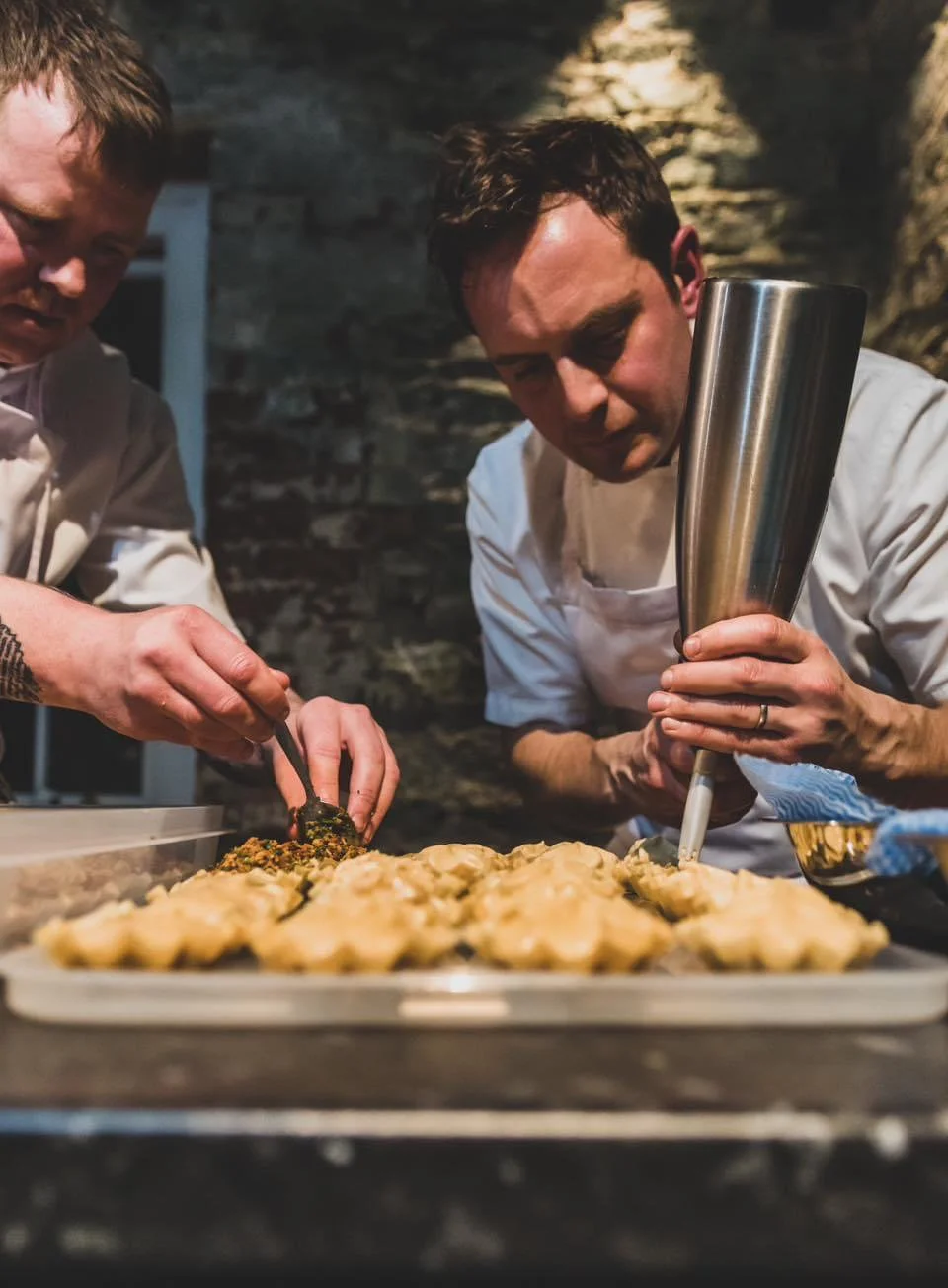 Two chefs garnish and prepare a tray of cooked food in a professional kitchen setting.
