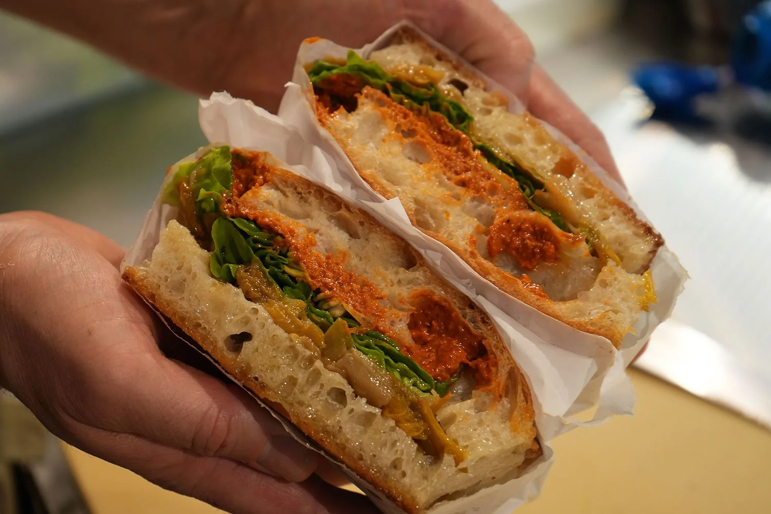 Close-up of a sandwich with green lettuce, melted cheese, red sauce, and crispy bread, held in a person's hand.