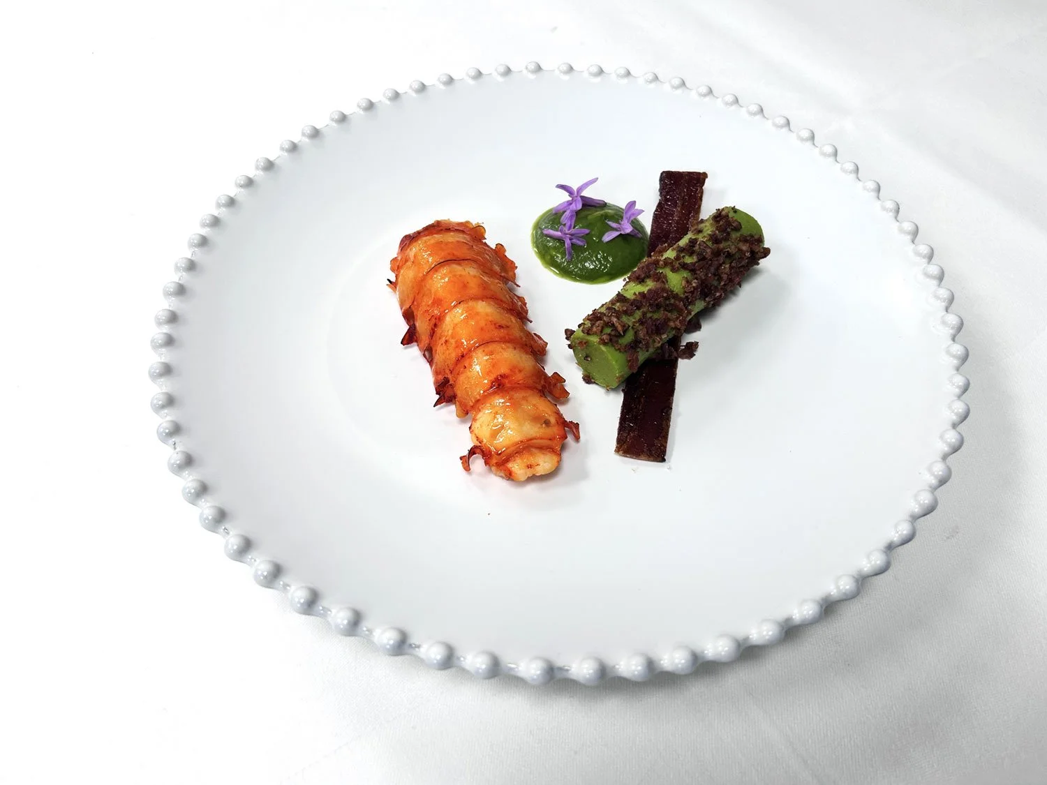 Gourmet plated dish with a lobster tail, a green mousse rolled with chocolate crumble, and a purple flower garnish on a white beaded-edge plate.