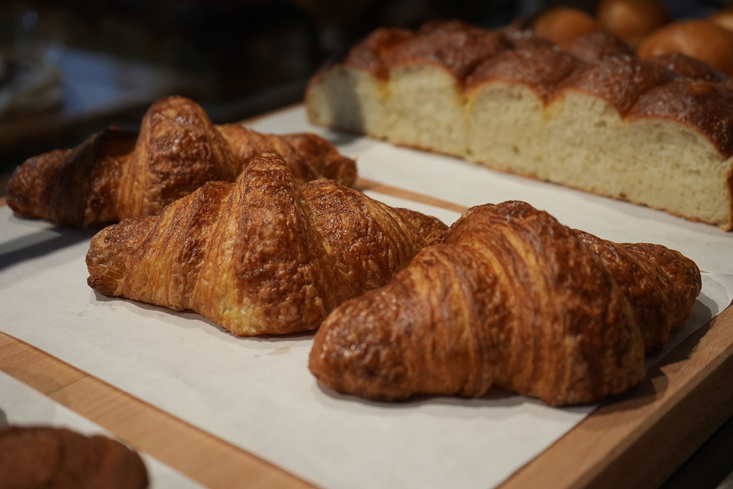 Close-up of three golden brown croissants and a loaf of sliced bread on a wooden tray with parchment paper.