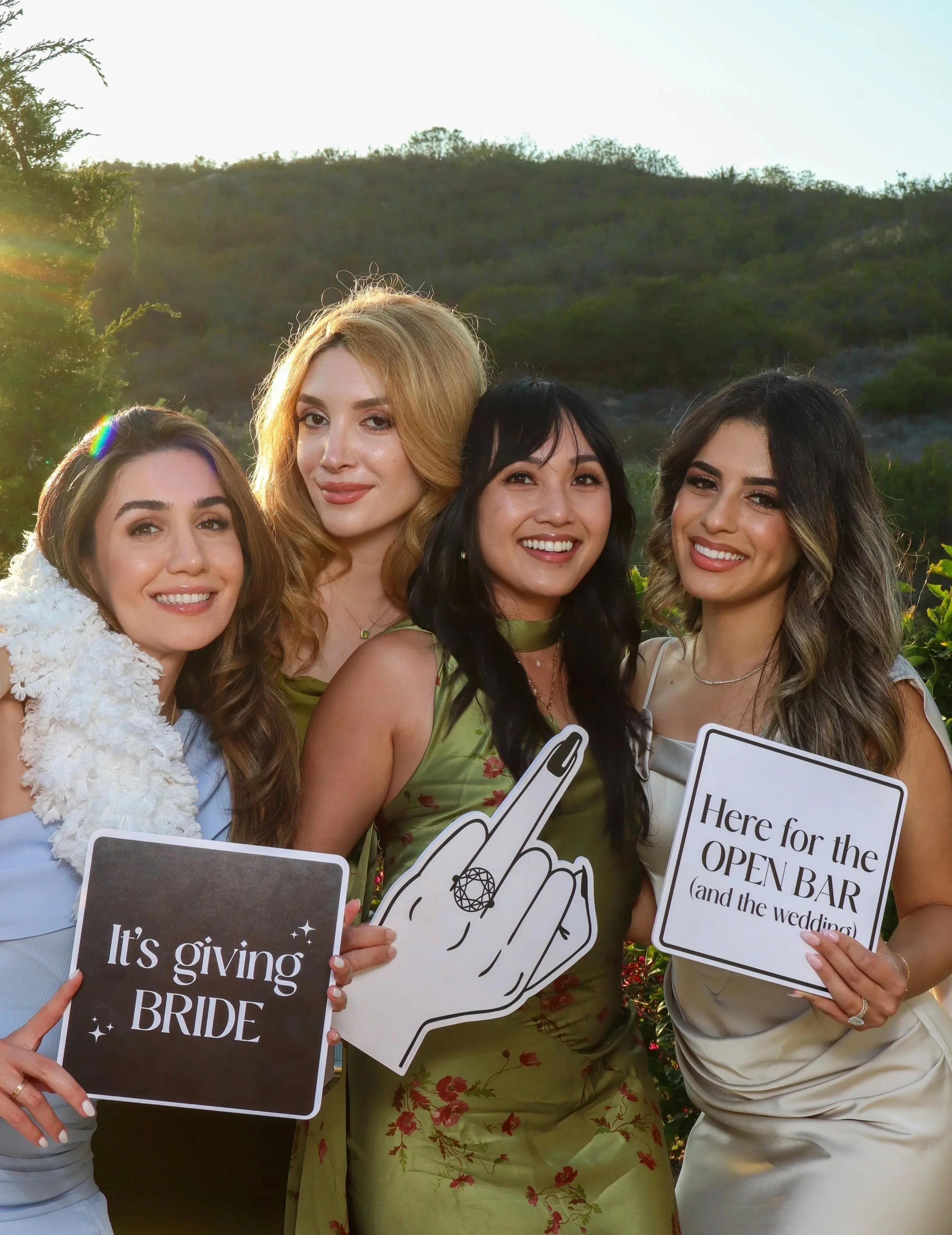 Group of women holding signs at a wedding event outdoors during the daytime, with a hill and greenery in the background.