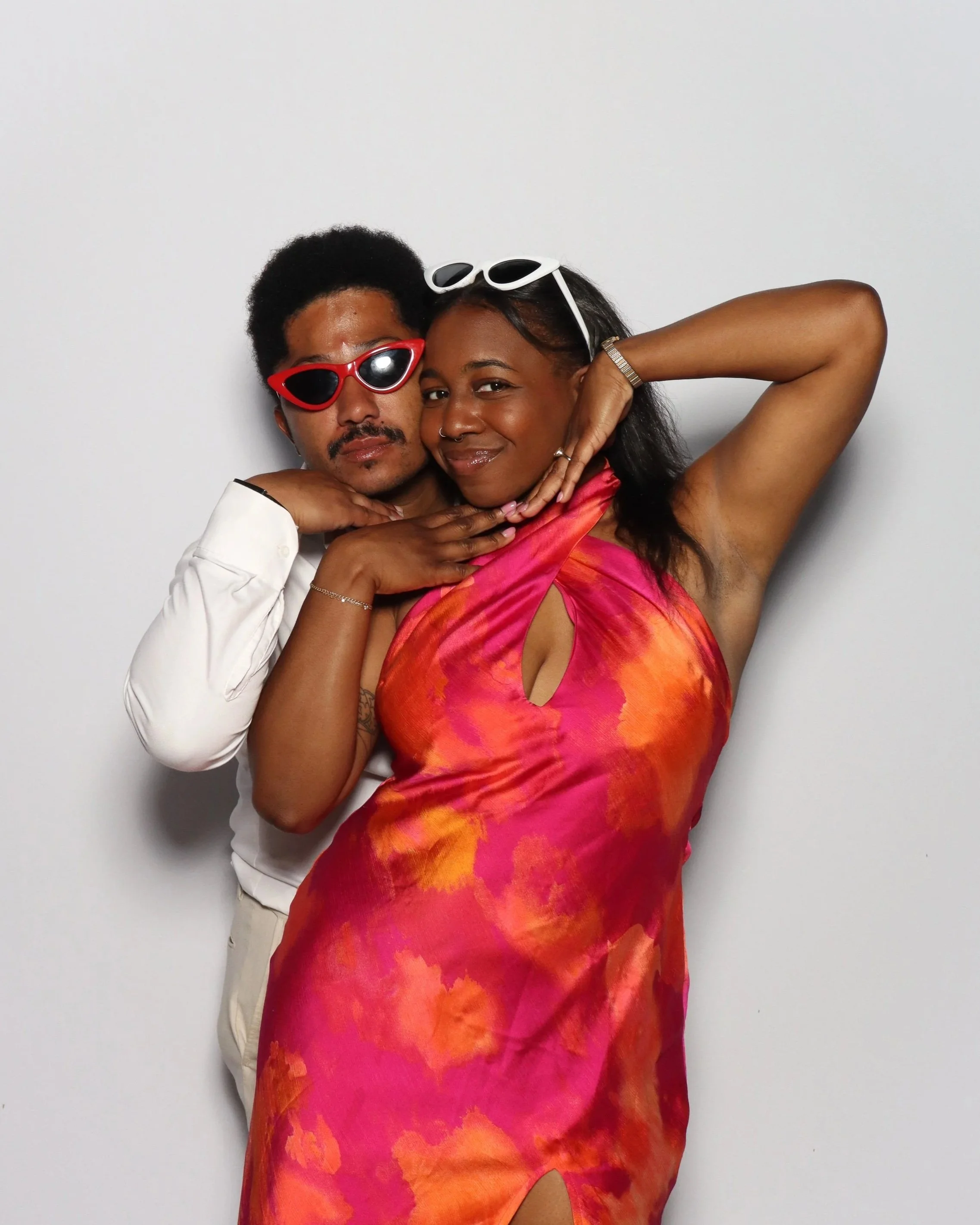 Two people posing closely together against a plain white background. The person on the left has short, curly black hair, is wearing red sunglasses, a white shirt, and has a mustache. The person on the right has long, dark hair, white sunglasses on th