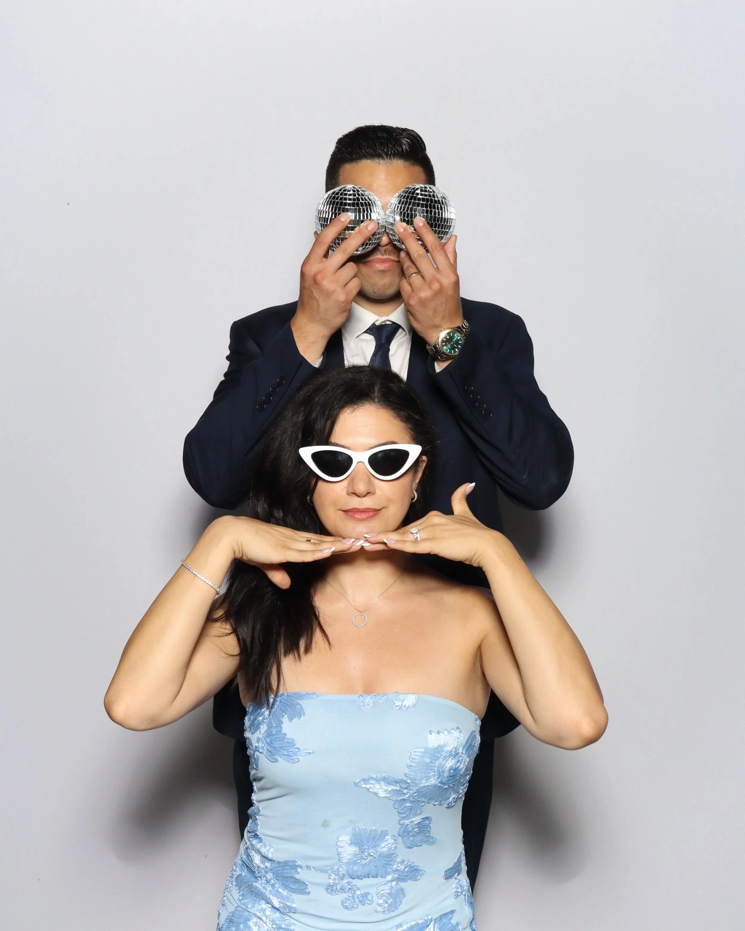 A man in a suit and tie holding two disco balls over his eyes, and a woman in sunglasses and a strapless dress posing with her hands under her chin.