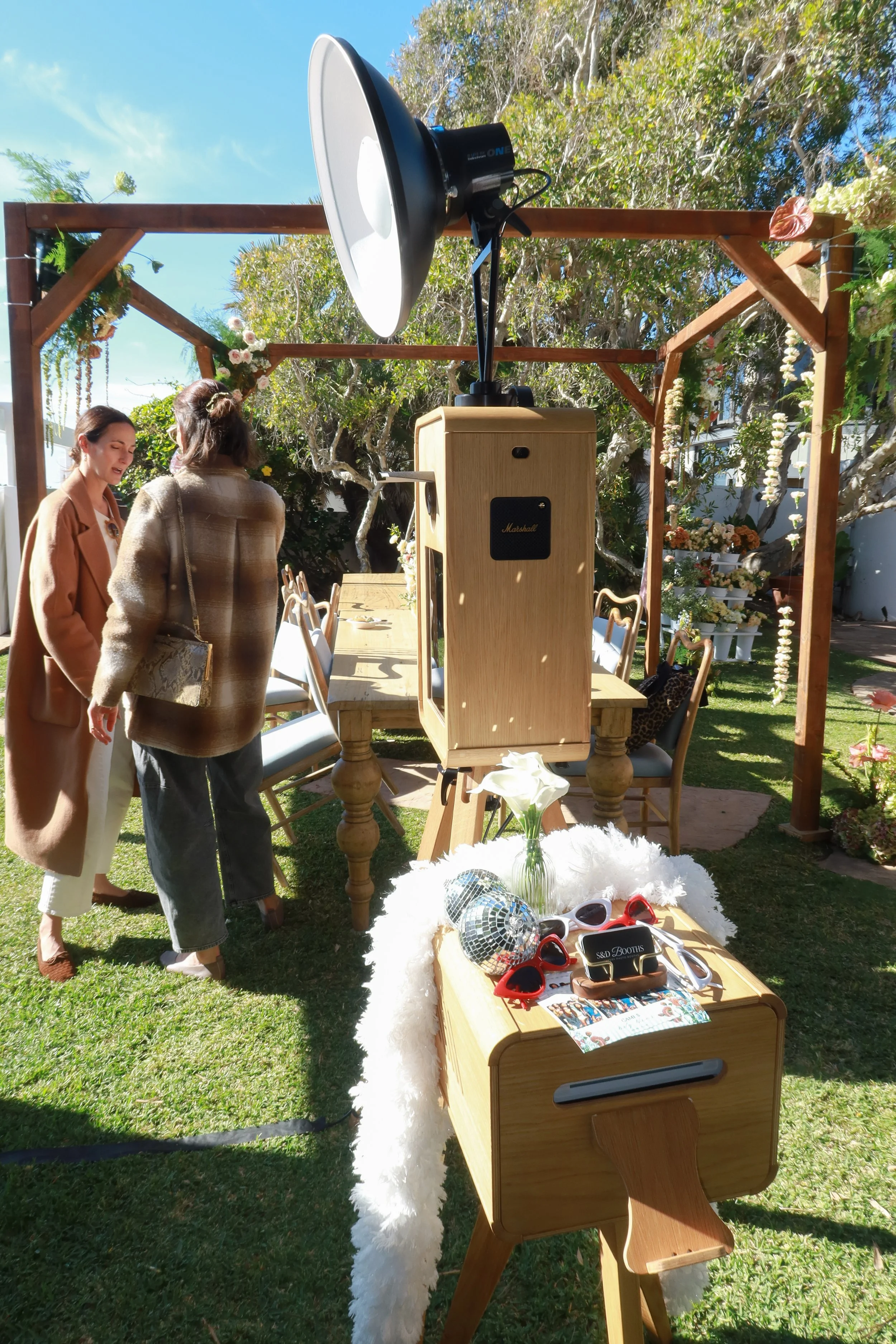 Outdoor photo of a photo booth setup at a garden party or wedding, featuring a large camera with a flash, decorative flowers, and props like sunglasses and disco balls on a table.