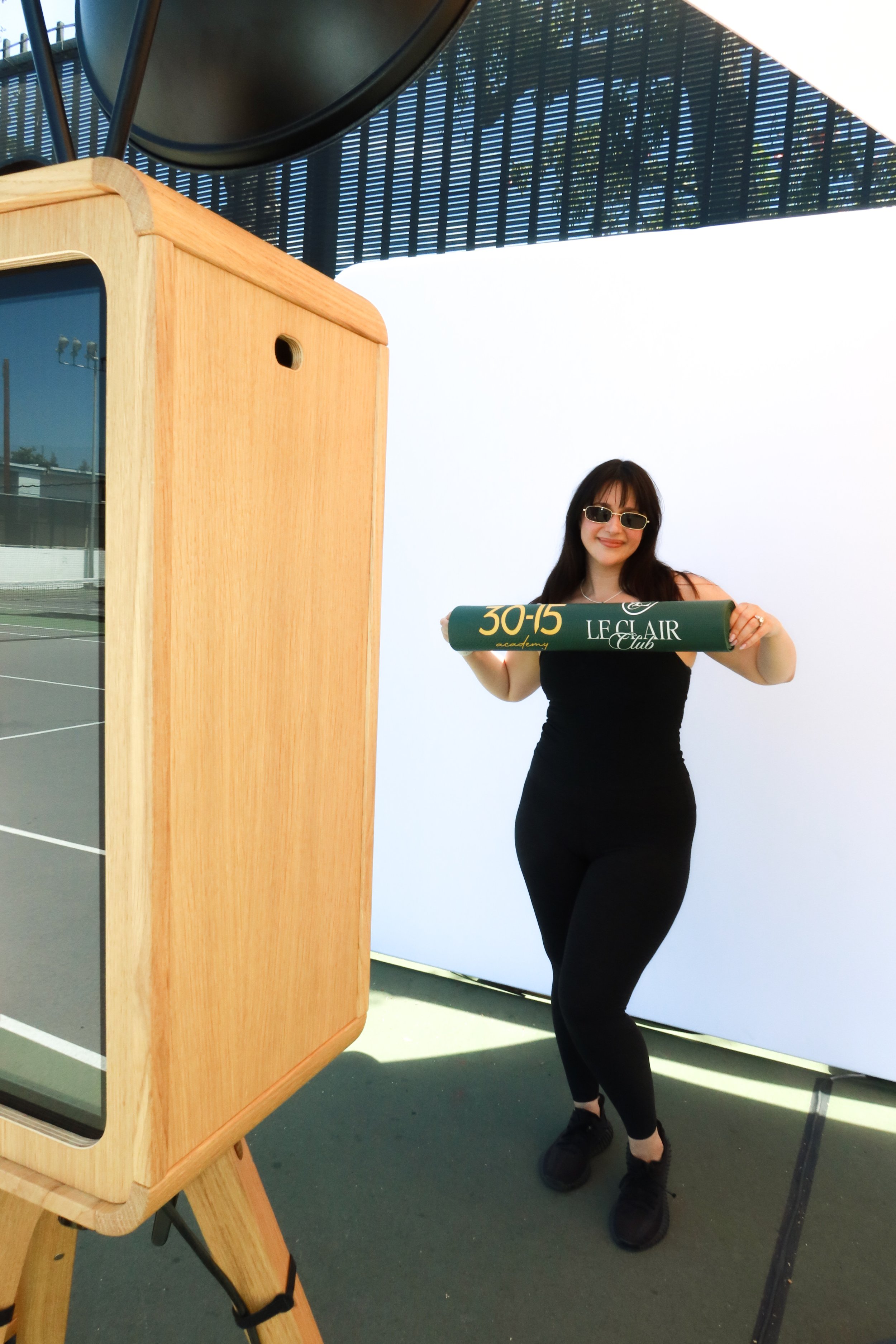 A woman wearing sunglasses and a black dress standing outdoors, holding a dark green sign that reads '30-15 LE CLAIR Club.' She is standing in front of a bright white background with a dark structure above.