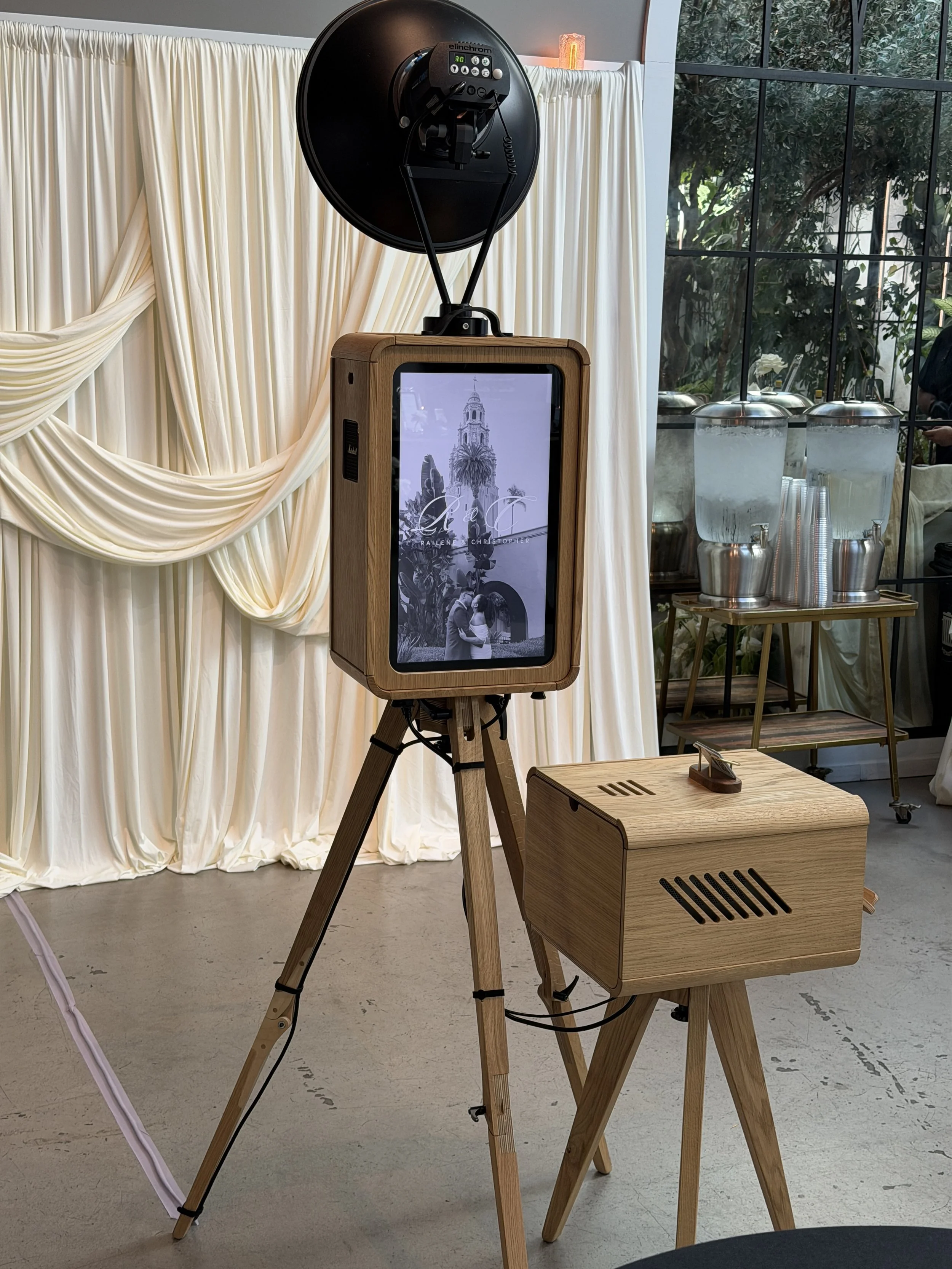 A photo booth setup with a wooden box containing a screen displaying a black-and-white image of a couple and an archway. The booth has a large round black reflector and is in a room with cream-colored draped curtains and large glass windows showing g