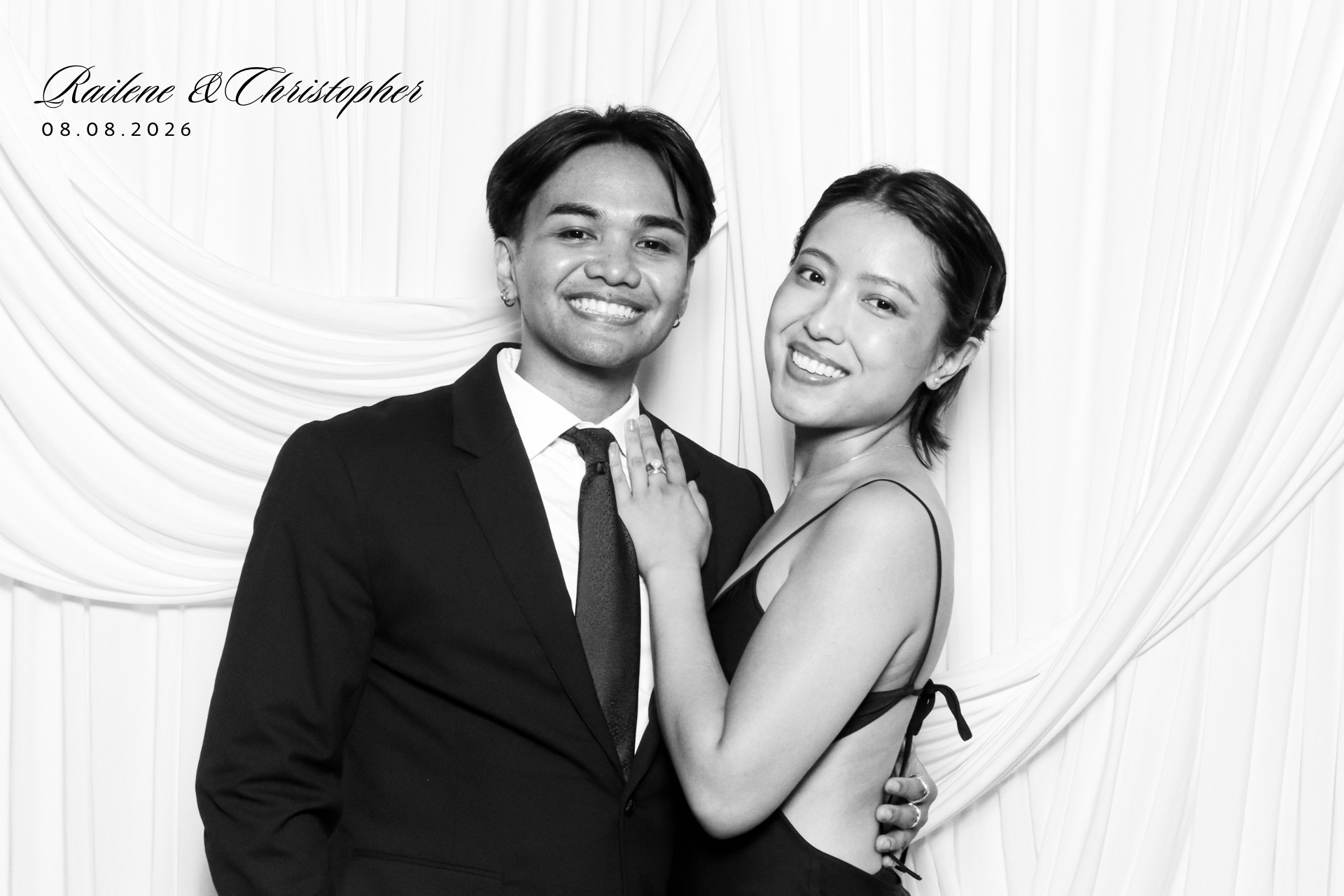 Black and white photo of a smiling couple at a wedding, with the woman showing her engagement ring. The man is wearing a suit and tie, and the woman is in a dress. They are standing in front of a draped backdrop. Text in the upper left corner reads "