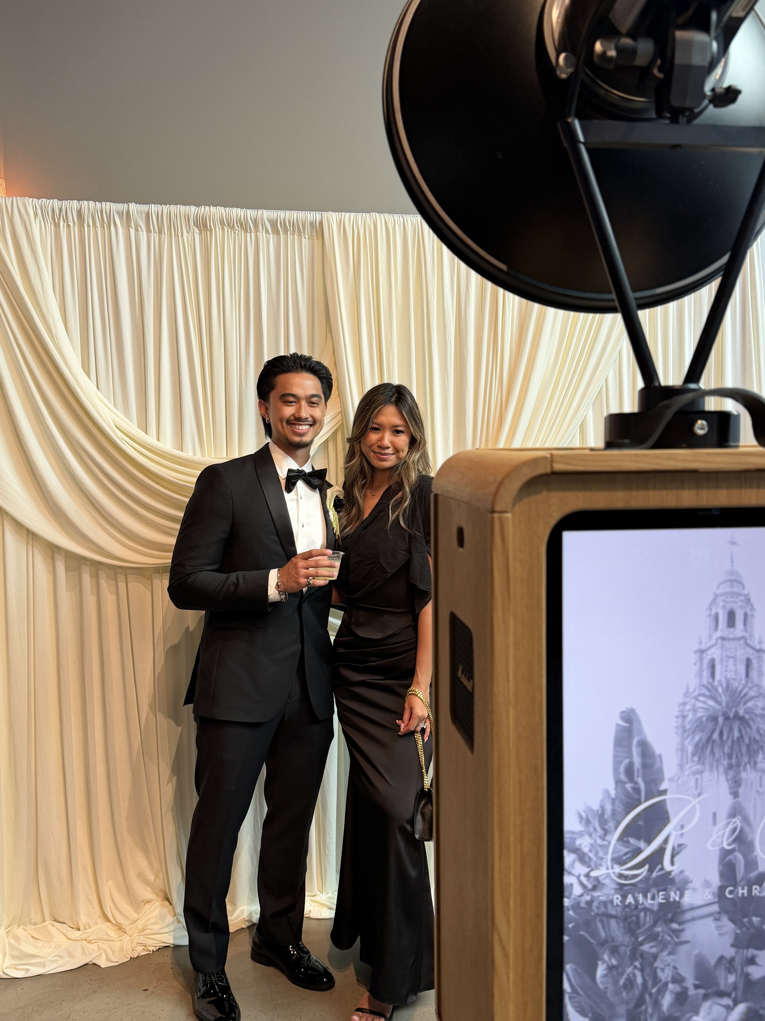 A man and woman dressed formally, standing in front of cream-colored draped curtains at an event or celebration. The man is wearing a tuxedo with a bow tie, and the woman is wearing a black dress holding a small handbag. They are smiling at the camer
