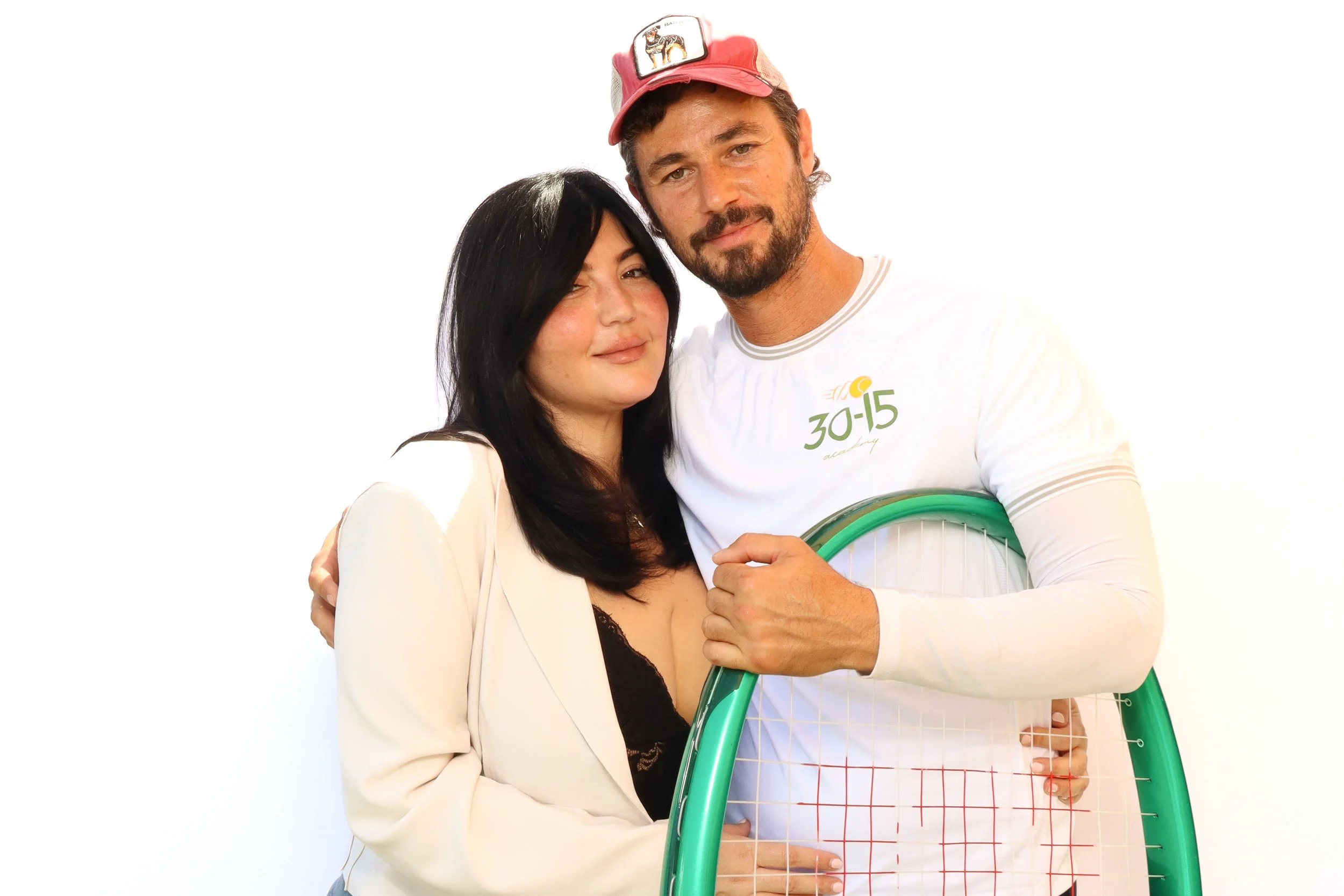 A man and woman stand close together against a plain white background. The man has a beard, light brown hair, and is wearing a pink cap and a white shirt with green and orange lettering. The woman has long black hair and is wearing a white blazer ove