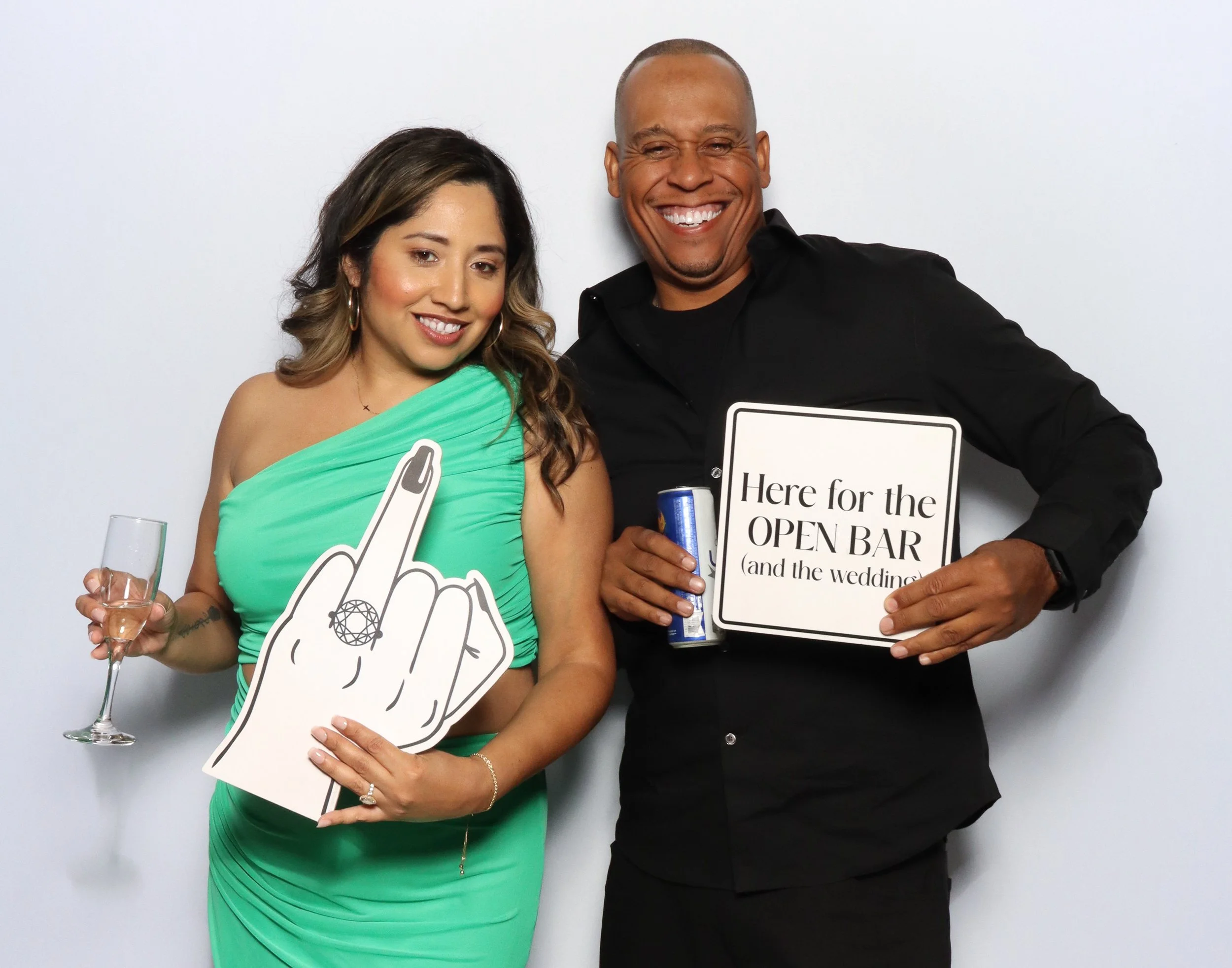 Two people smiling at a photo booth, holding signs and drinks, with a white background.