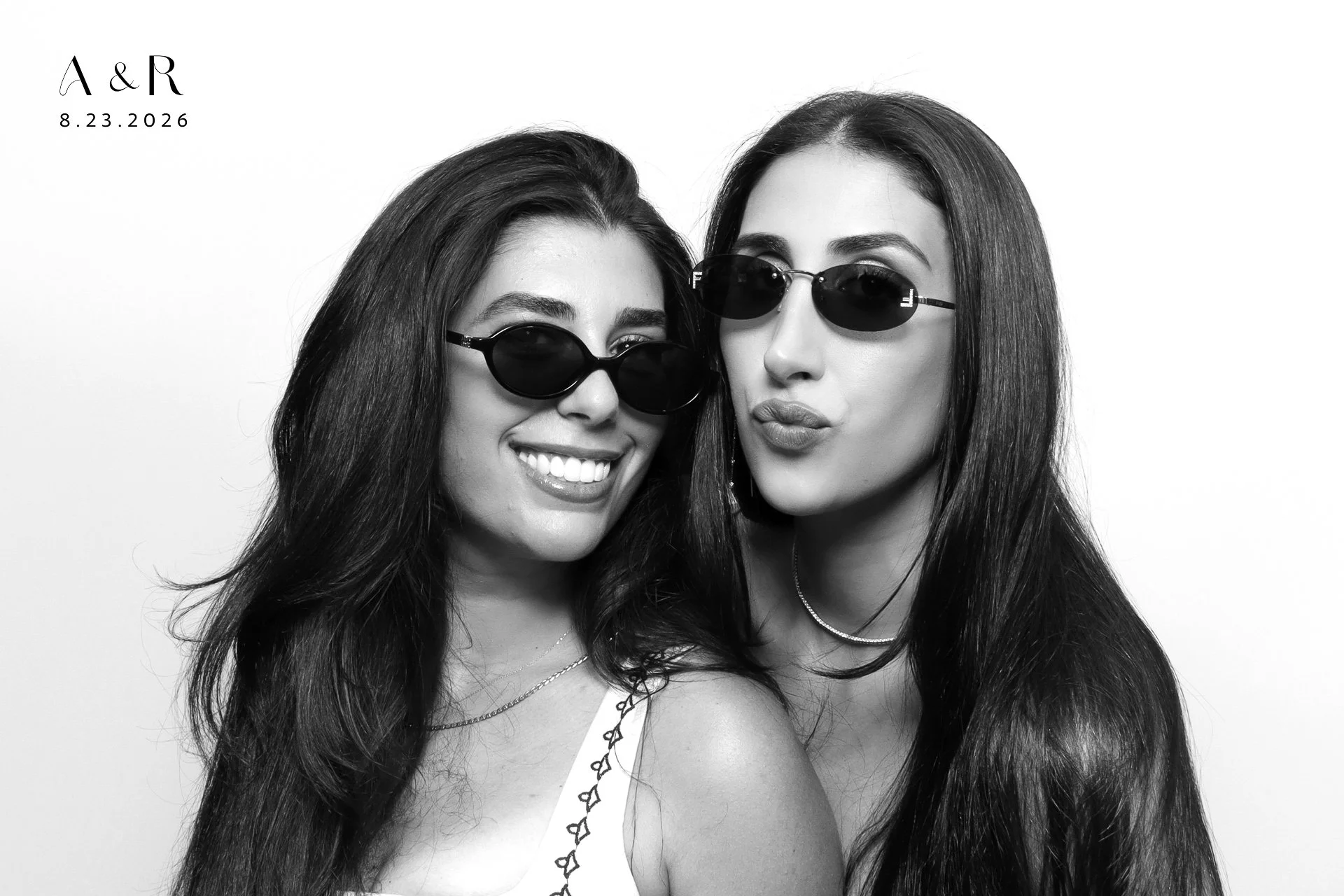 Black and white photo of two young women wearing sunglasses, smiling and leaning close to each other against a plain background. Text in the top left corner reads "A & R" with a date of "8.23.2026" below.