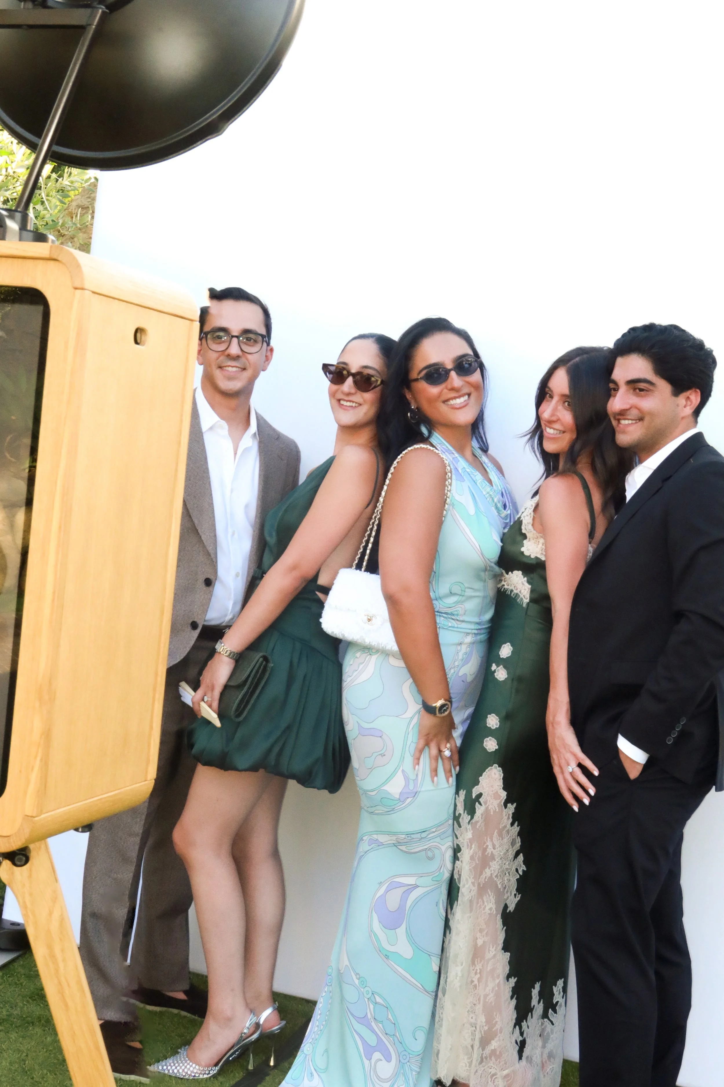 Five people dressed in formal and semi-formal attire, posing together for a photo during an outdoor event, with a vintage photo booth on the left side of the image.