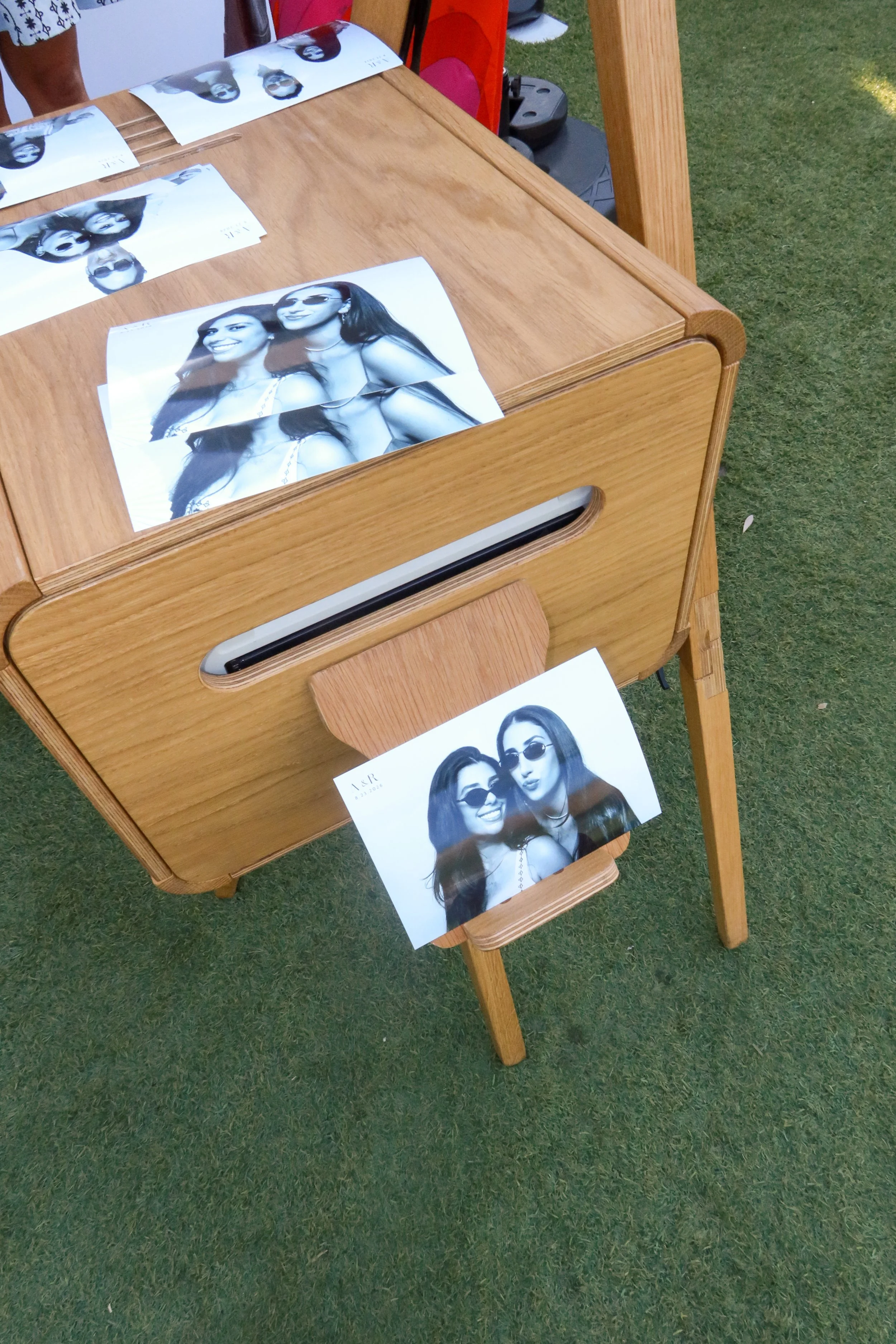 Photographs of two women in black and white at an outdoor event, displayed on a wooden table with a slot for papers, on green grass.