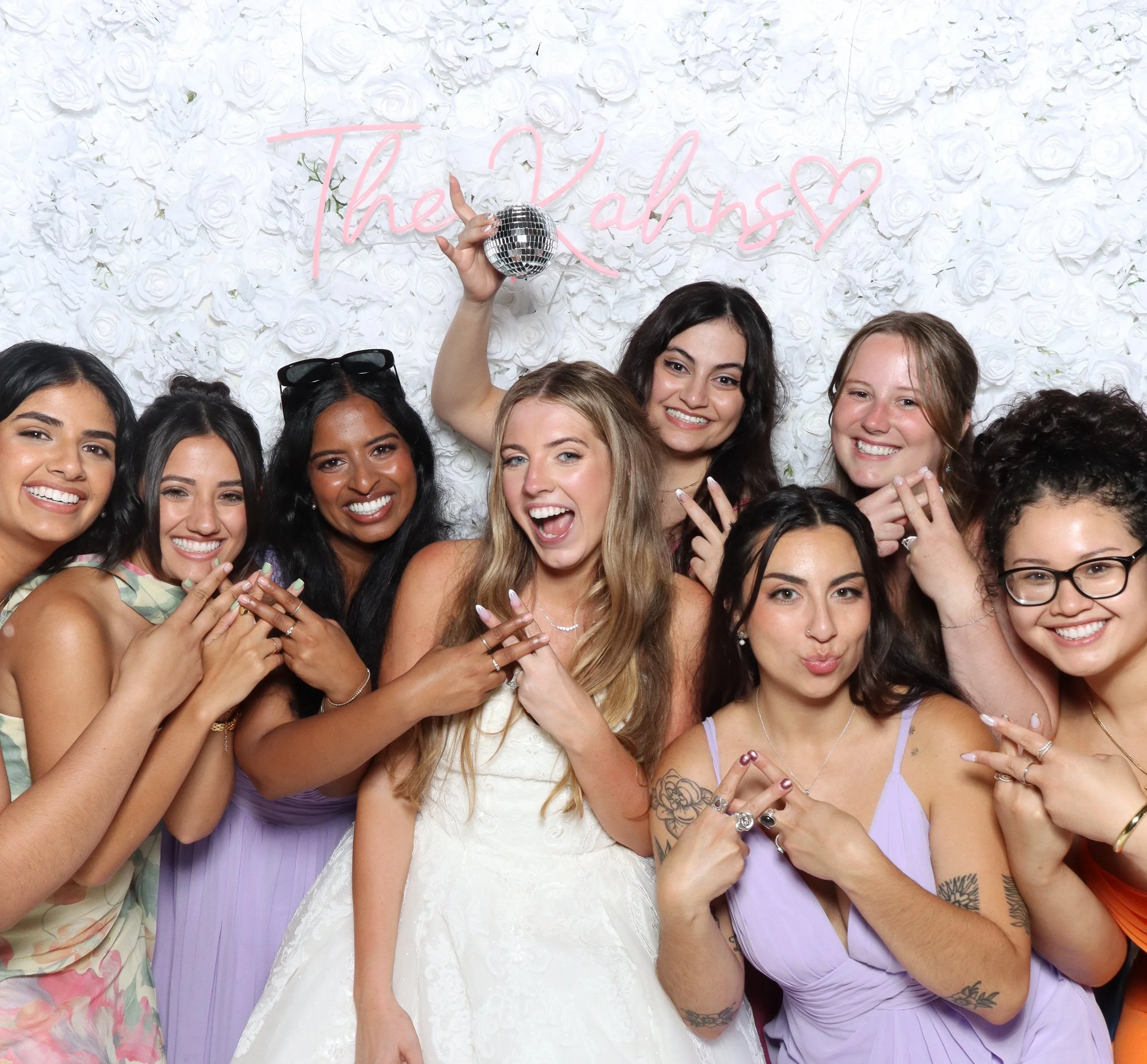 Group of diverse women smiling, posing, and celebrating in front of a white floral backdrop with a pink neon sign reading 'The Ams' and a small disco ball.