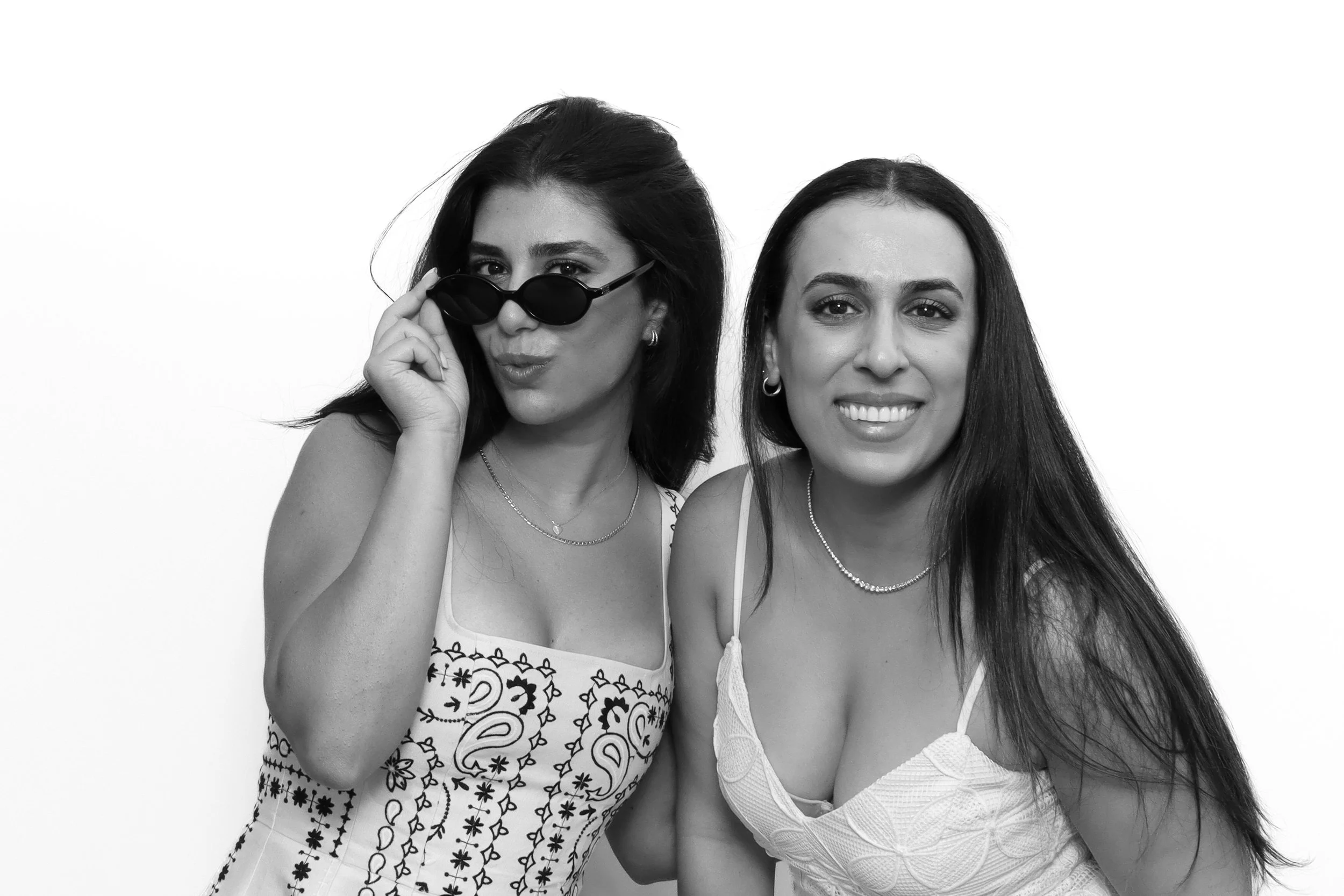 Two women with dark hair, smiling, dressed in summer clothing, standing close together against a plain white background. The woman on the left has dark hair, wears sunglasses, and a patterned dress. The woman on the right has her hair styled down and