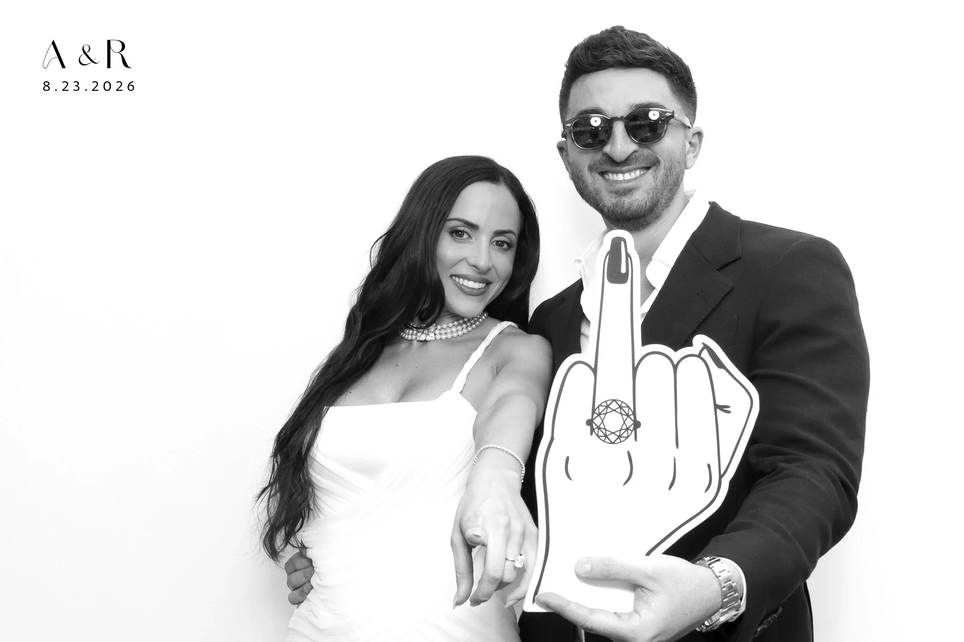 A smiling couple, a woman with long dark hair wearing a white dress and pearl necklace, and a man with short hair, beard, sunglasses, and a suit, holding a cartoon cutout of a hand with a ringed middle finger.