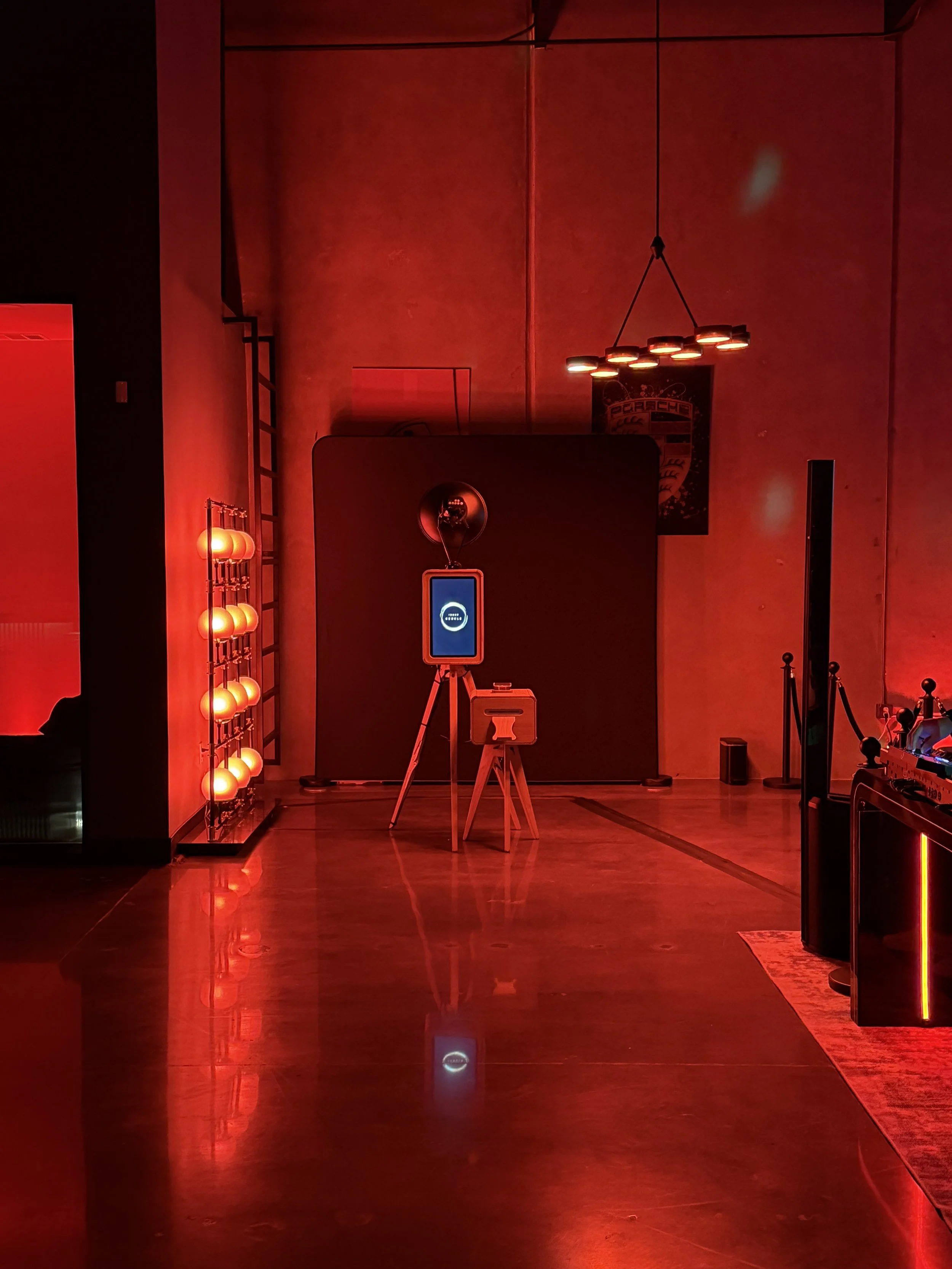 A dimly lit room with red lighting, featuring a modern chandelier hanging from the ceiling, a small traffic light selfie stand, a vintage radio, and a Porsche poster on the wall.