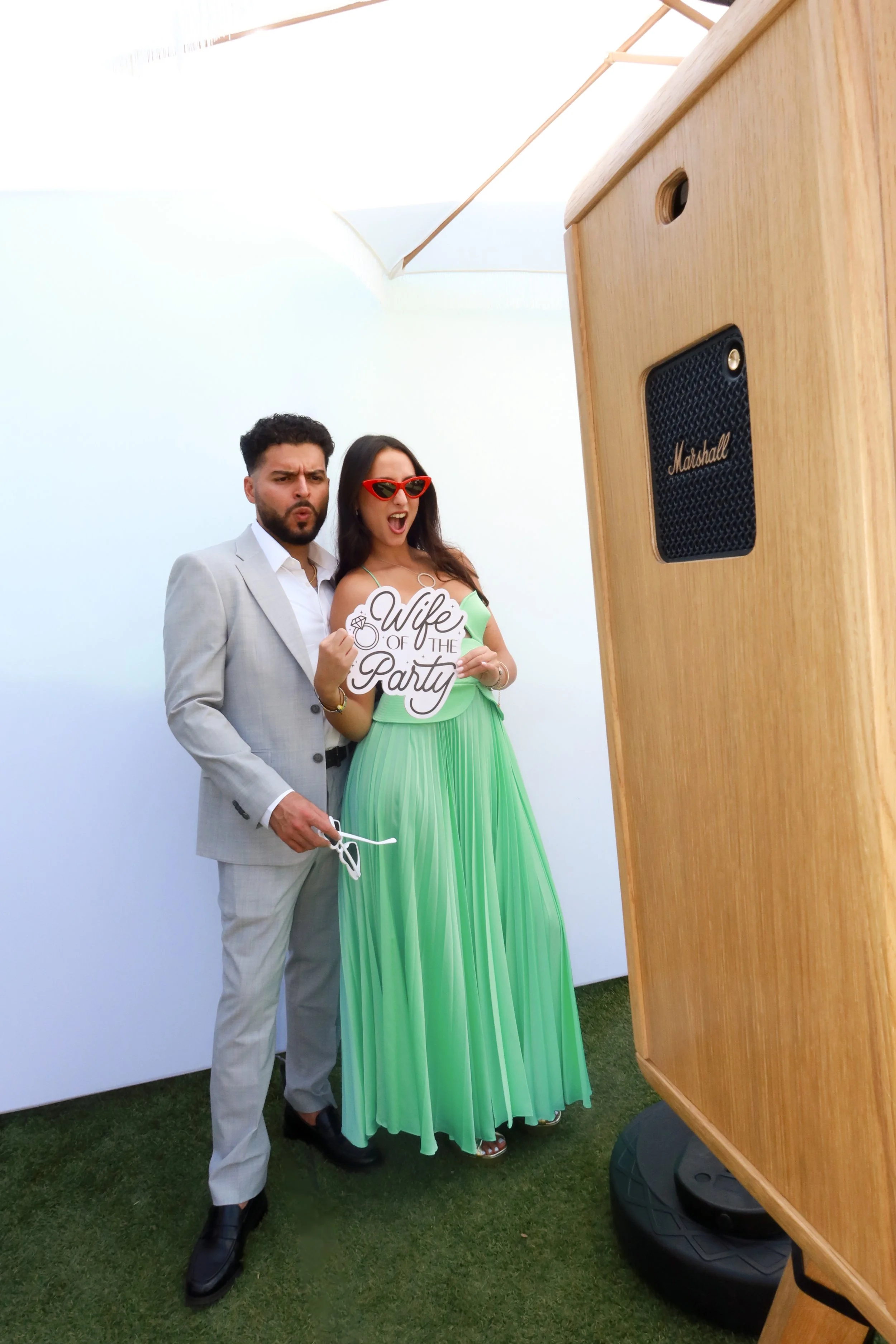 A man and a woman posing at a party, the woman holding a sign that says 'Wife of the Party'. They are standing outdoors near a large speaker and a white backdrop, with a grassy ground beneath them.