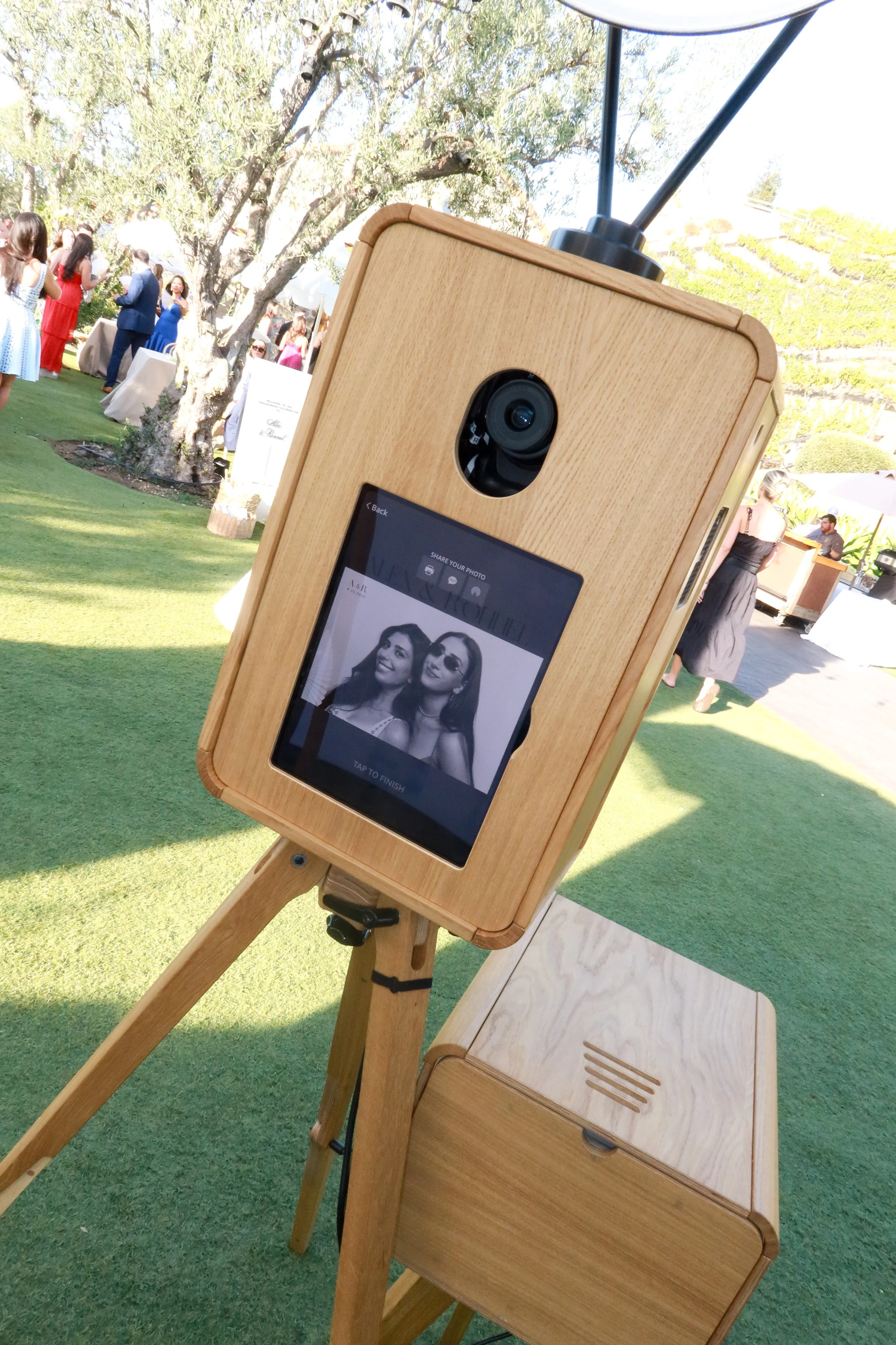 A wooden photo booth with a digital screen and camera on a stand, set outdoors at a gathering with people in the background, trees, grass, and tables.