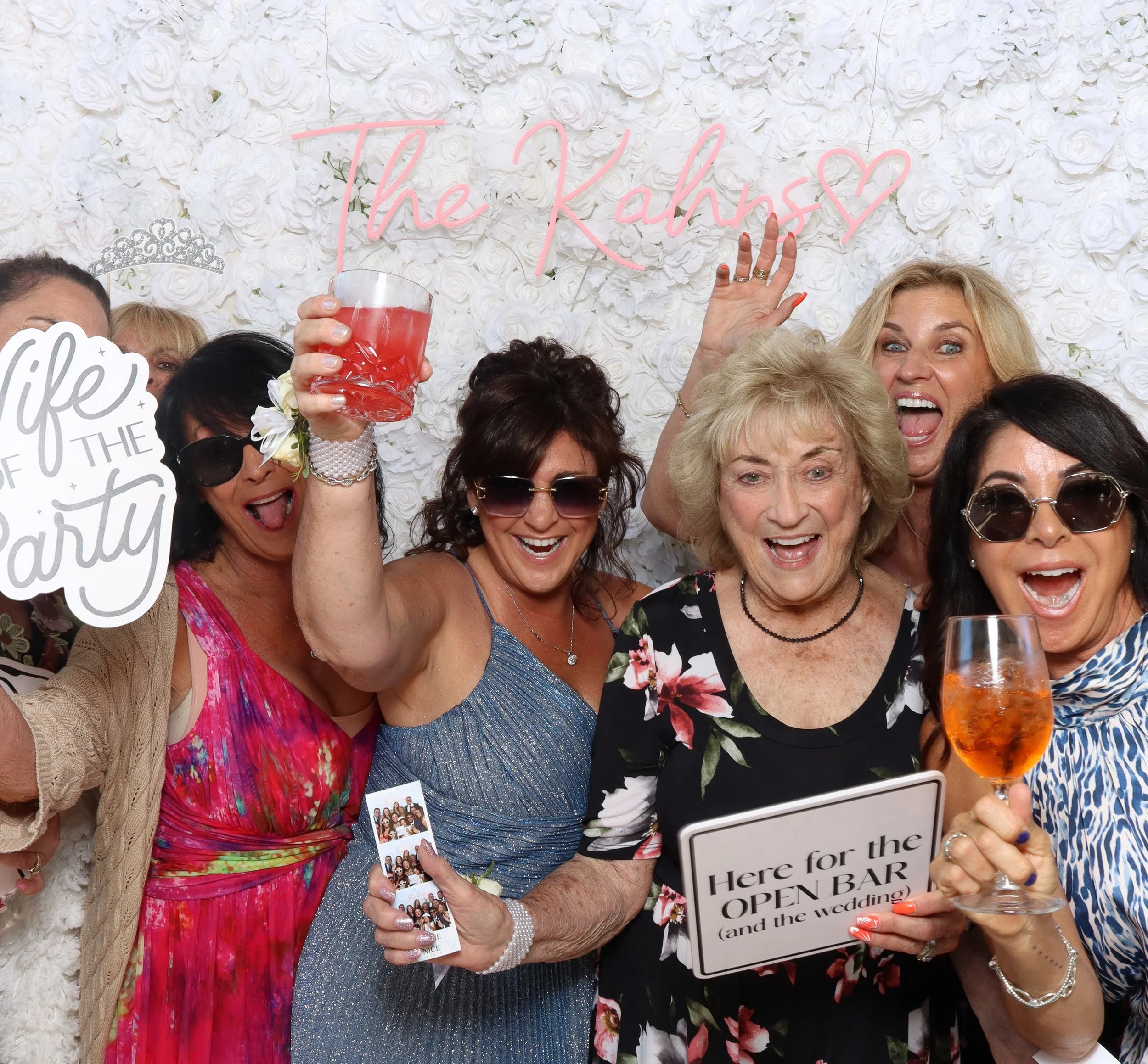 Celebration with women wearing tiaras and holding drinks, in front of a white floral backdrop with pink neon sign reading 'the Kahn's'.