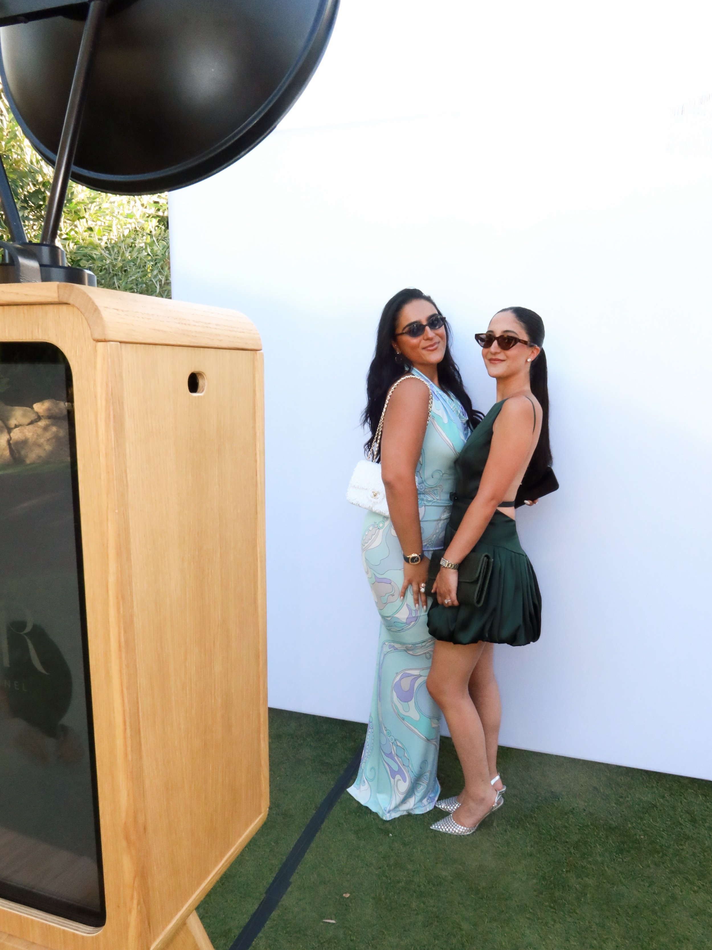 Two women wearing sunglasses and elegant dresses posing and smiling next to a photo booth at an outdoor event.