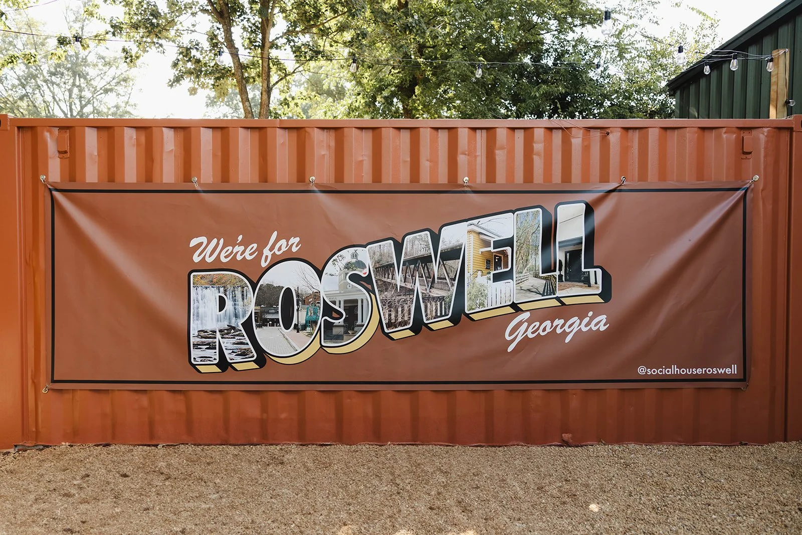 A large brown banner on a rusty metal fence reads 'We're for ROsWELL Georgia' with the word 'ROSWELL' filled with images of local landmarks, trees, and buildings, and the social media handle '@socialhouseroswell' in the lower right corner.